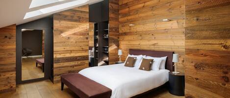 A modern bedroom with a skylight, wooden walls, a large bed with pillows, a bench, and a nightstand with a lamp.