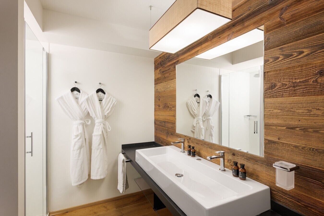 A modern bathroom with a large mirror, wooden wall paneling, a white sink, and two white robes hanging on the wall.