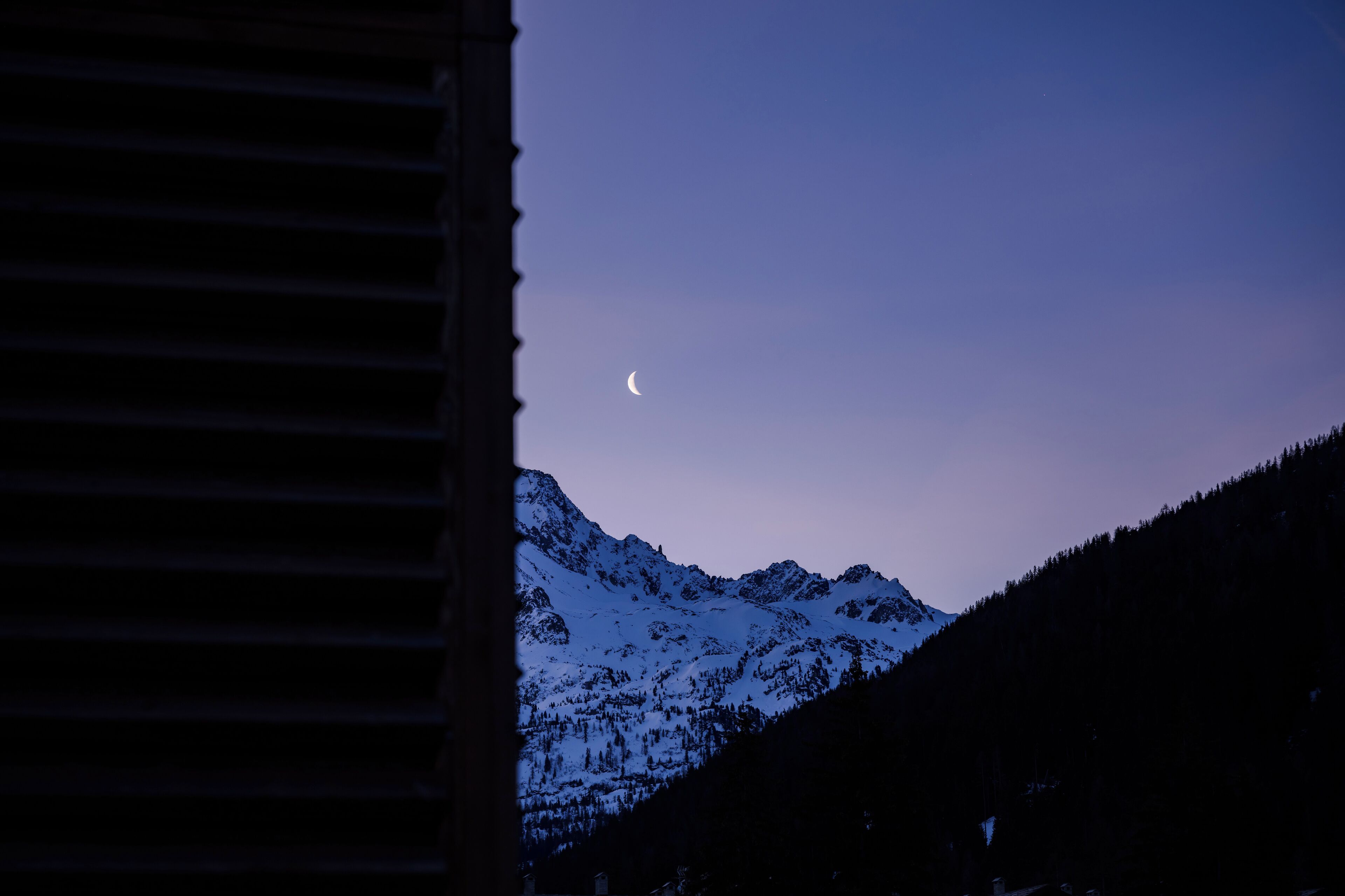 Catena montuosa con una luna crescente nel cielo, vista da una finestra con persiane orizzontali.