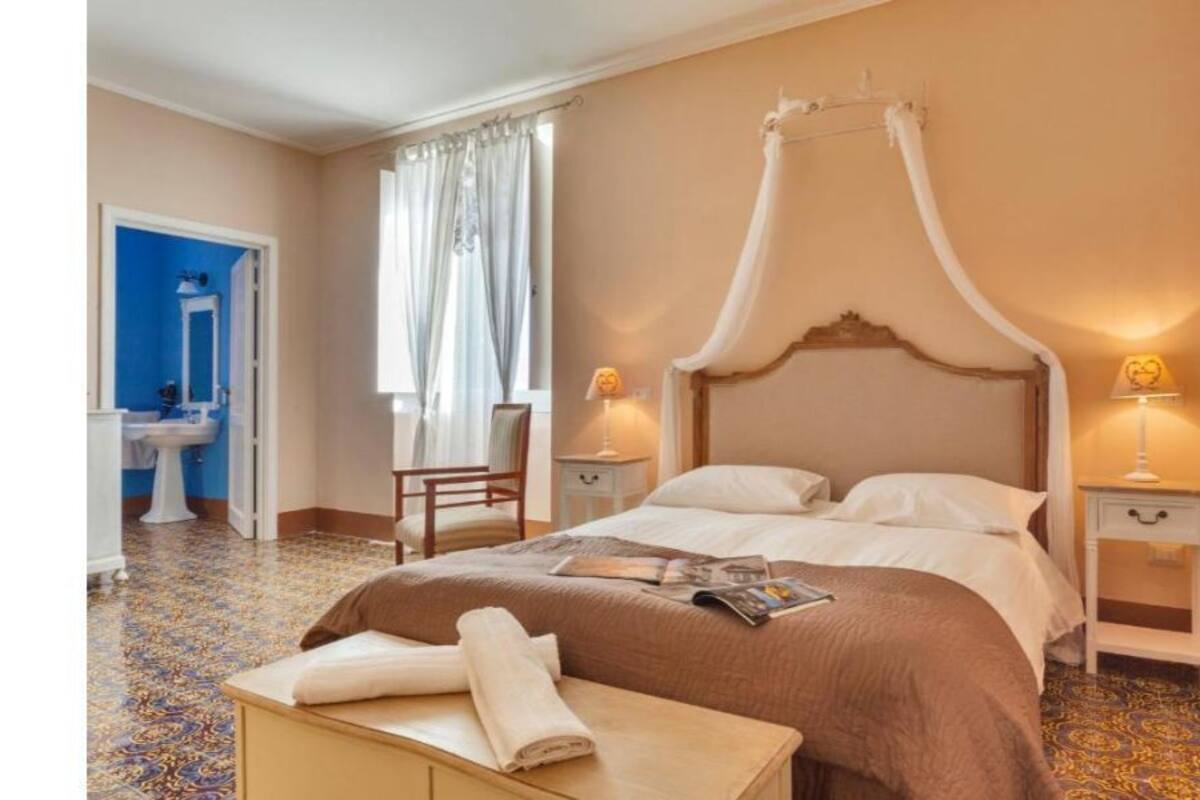 Een hotelkamer met een hemelbed, een zithoek met een stoel en een klein tafeltje, een nachtkastje met een lamp, en een badkamer zichtbaar door een geopende deur.