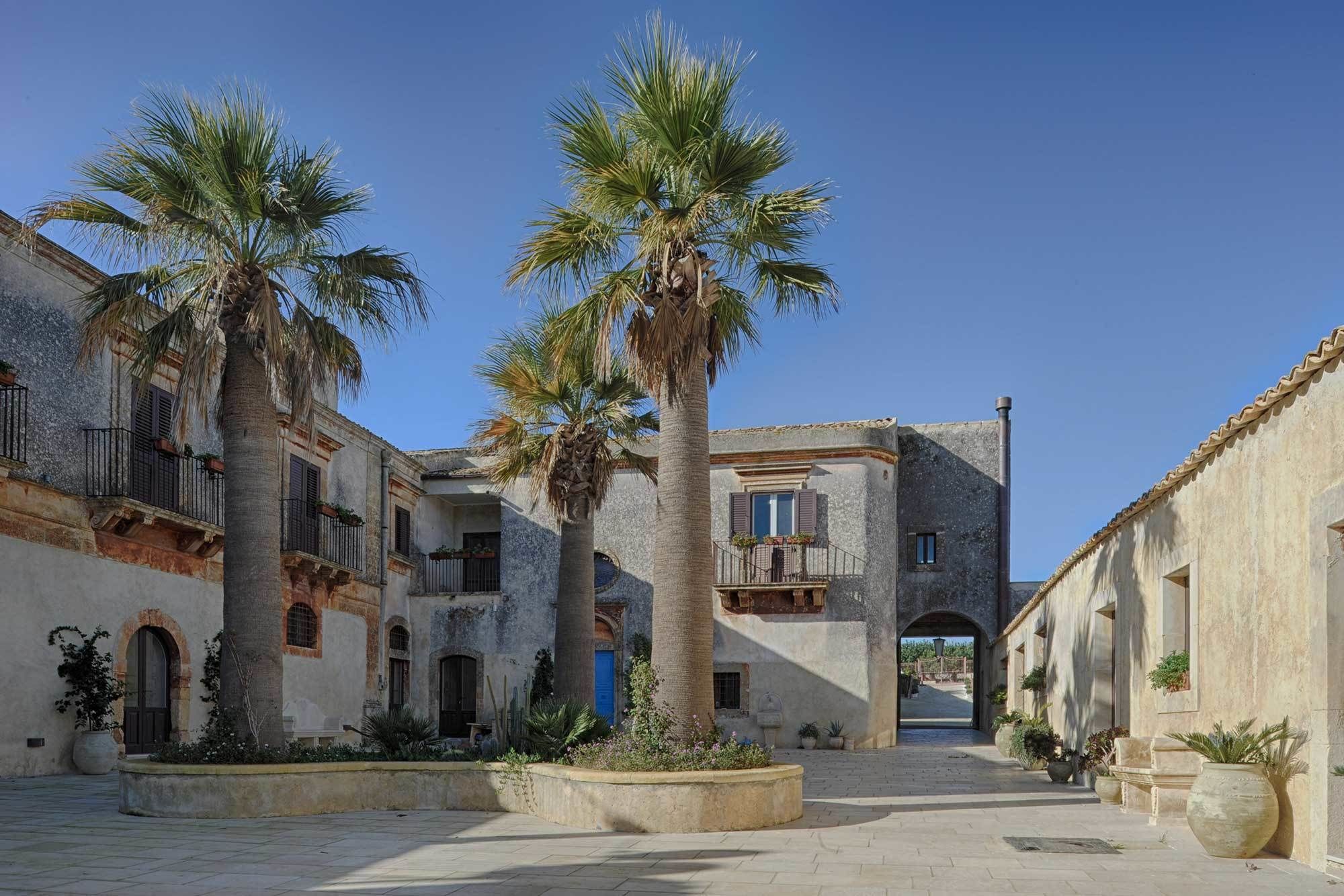 A courtyard with palm trees, stone buildings, and a clear blue sky.