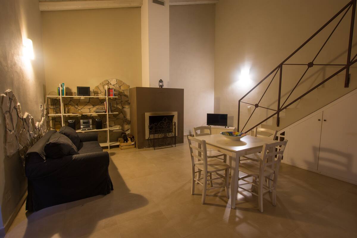 Een woonkamer met een open haard, een bank, een eettafel met stoelen en een trap.