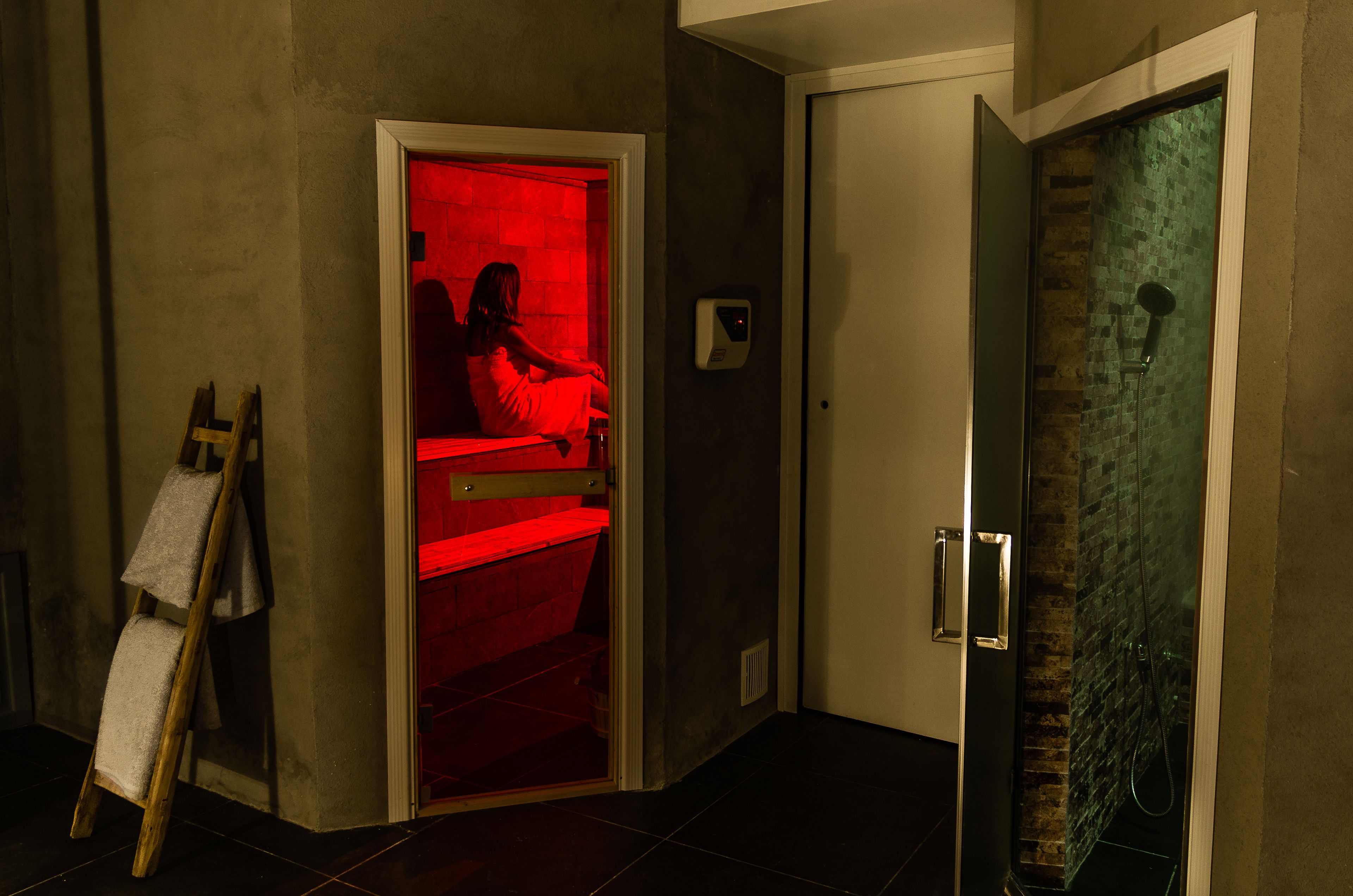 Ein Zimmer mit roter Beleuchtung in einer Ecke, eine Dusche mit Glastür und eine an der Wand lehnende Holzleiter.