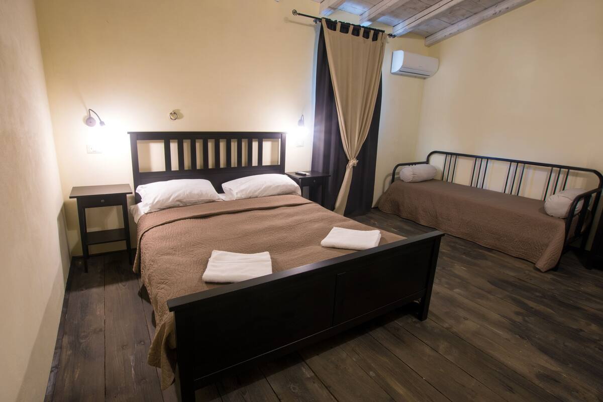 Een slaapkamer met een houten bed, een nachtkastje, een bankje en een gordijn.