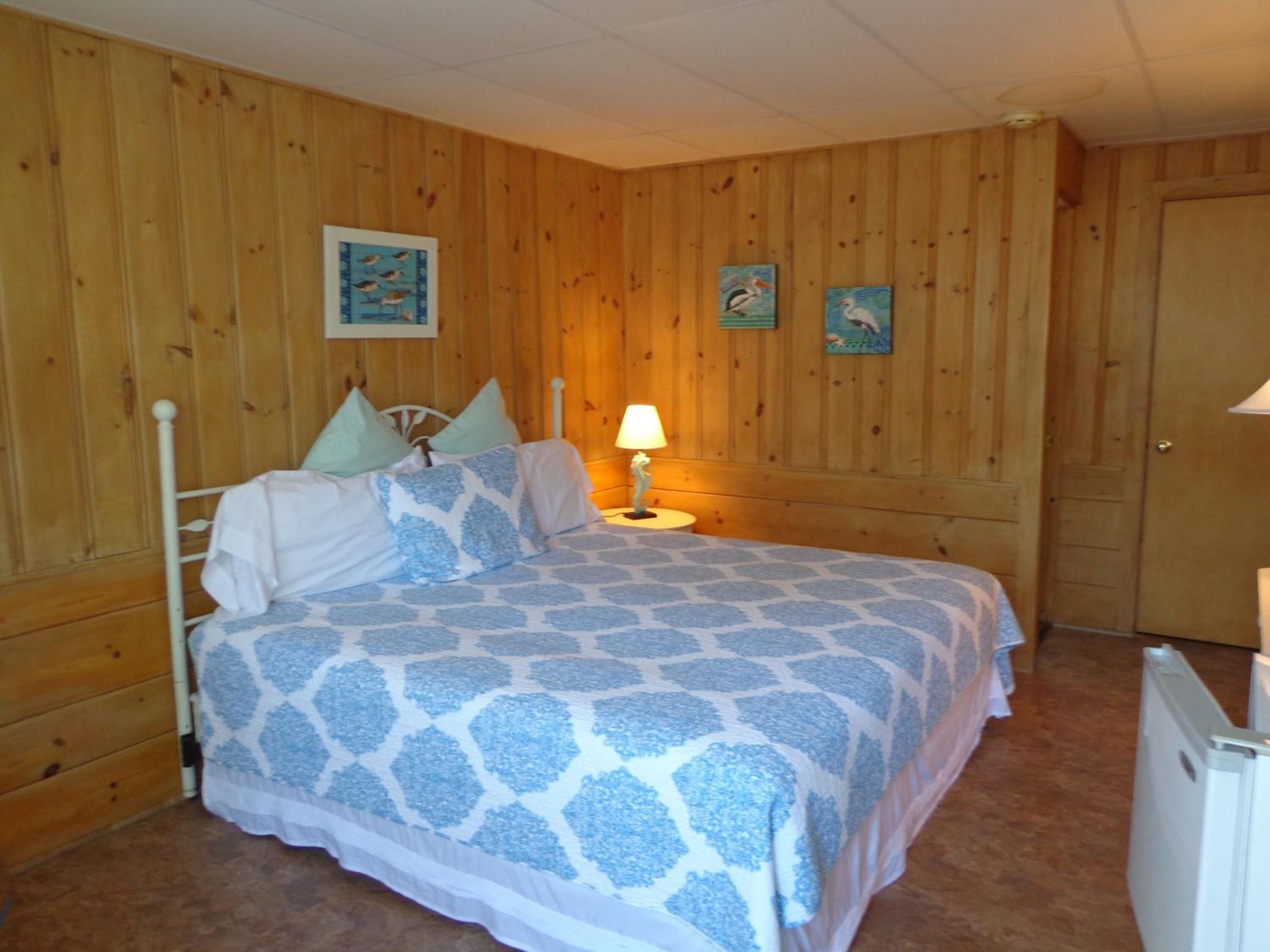 Un dormitorio con un revestimiento de madera, una cama con colcha estampada, una mesita de noche con lámpara y dos cuadros colgados en la pared.