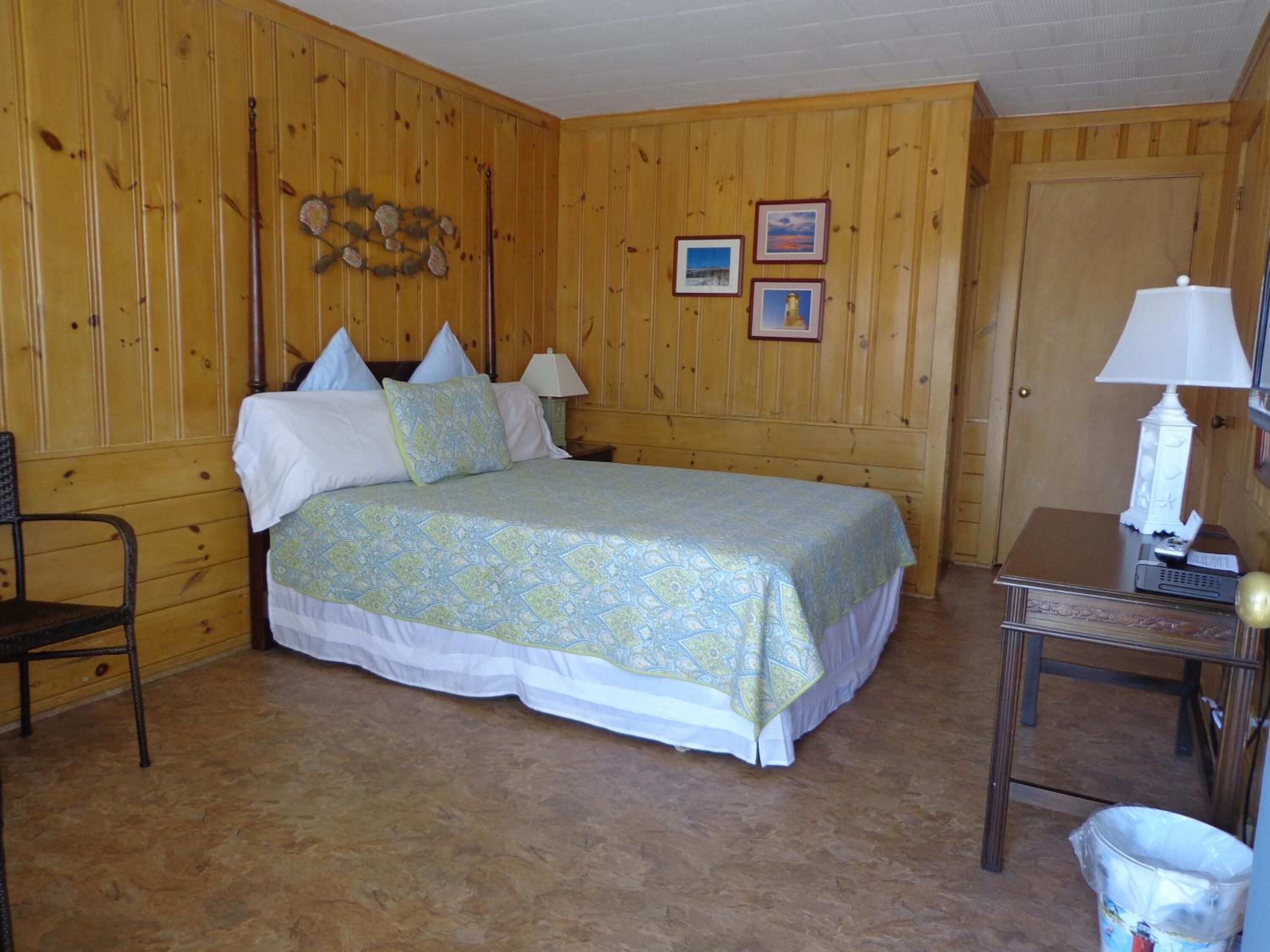 Una habitación con paneles de madera, una cama, una silla, una mesa con una lámpara y un cubo de basura.