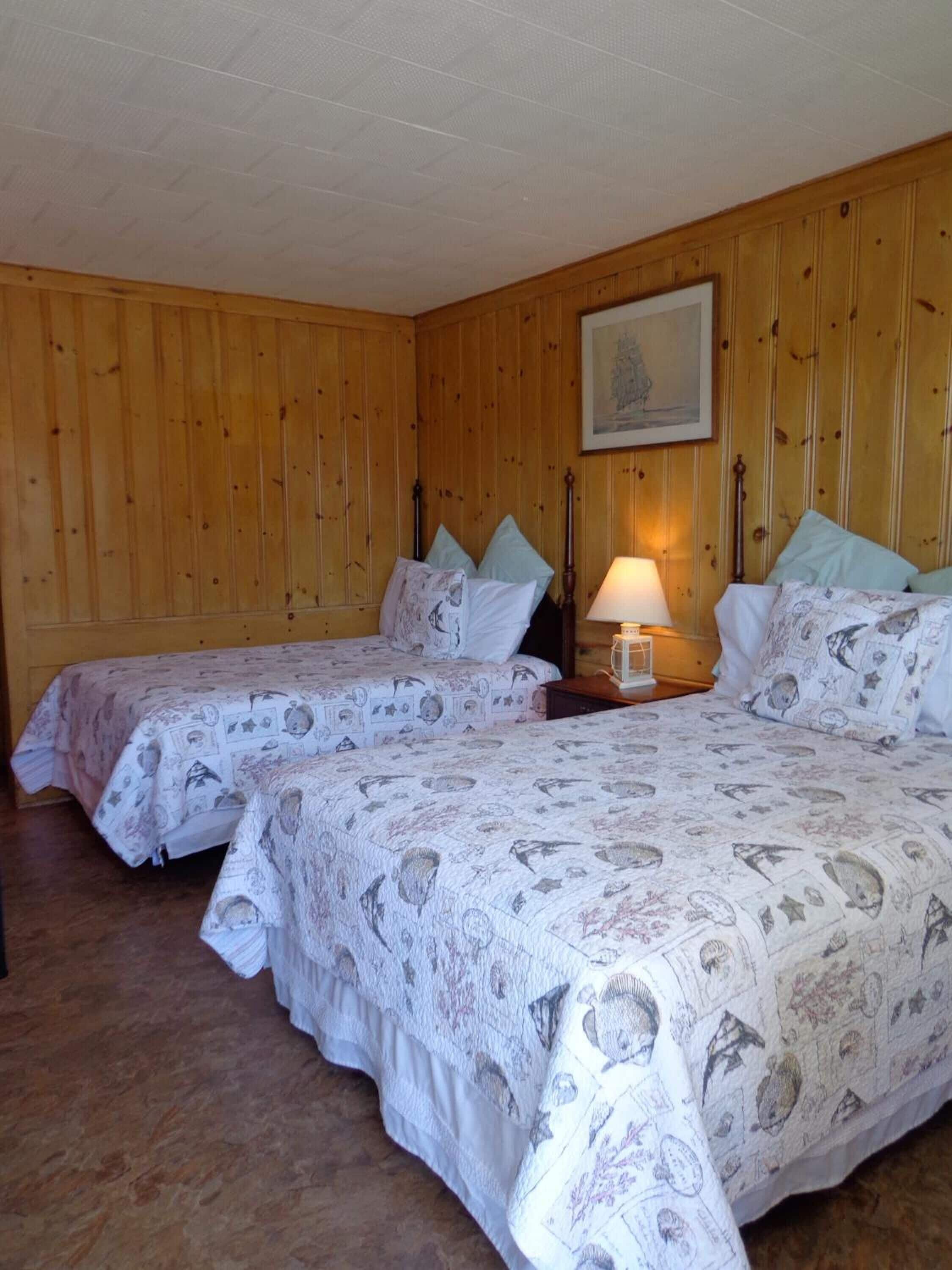 A room with two beds, wooden walls, a painting on the wall, and a nightstand with a lamp.