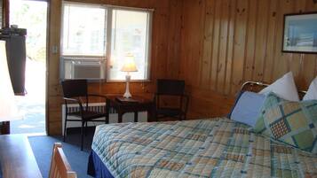 A wooden-paneled room with a bed, two chairs, a table, and a window with blinds.