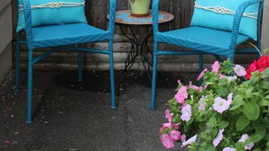 Una cerca de madera con un letrero que dice 'WELCOME', dos sillas de mimbre azules con cojines, una mesita con una planta en maceta y flores.