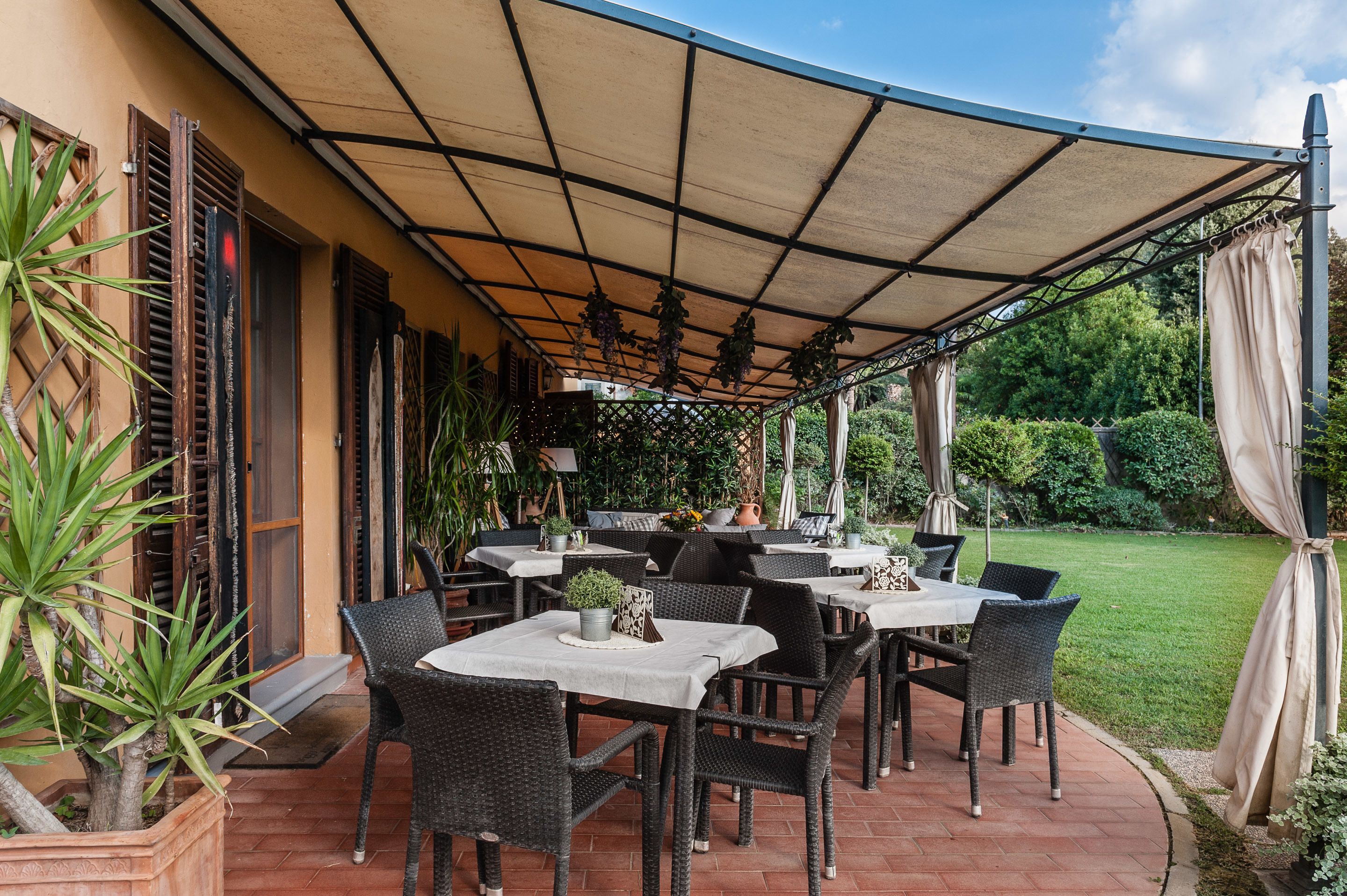 Une terrasse extérieure avec des tables et des chaises, entourée de plantes en pot et dotée d’un espace abrité.