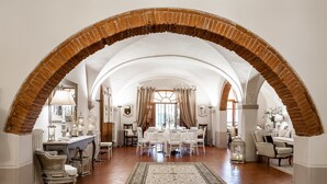 Una habitación amplia con un alto techo abovedado de ladrillo, un área de comedor central con mesa y sillas blancas, y una alfombra estampada en el suelo.