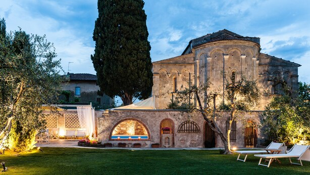 Edificio histórico con patio, destacando un gran árbol, asientos al aire libre y una entrada bien iluminada.