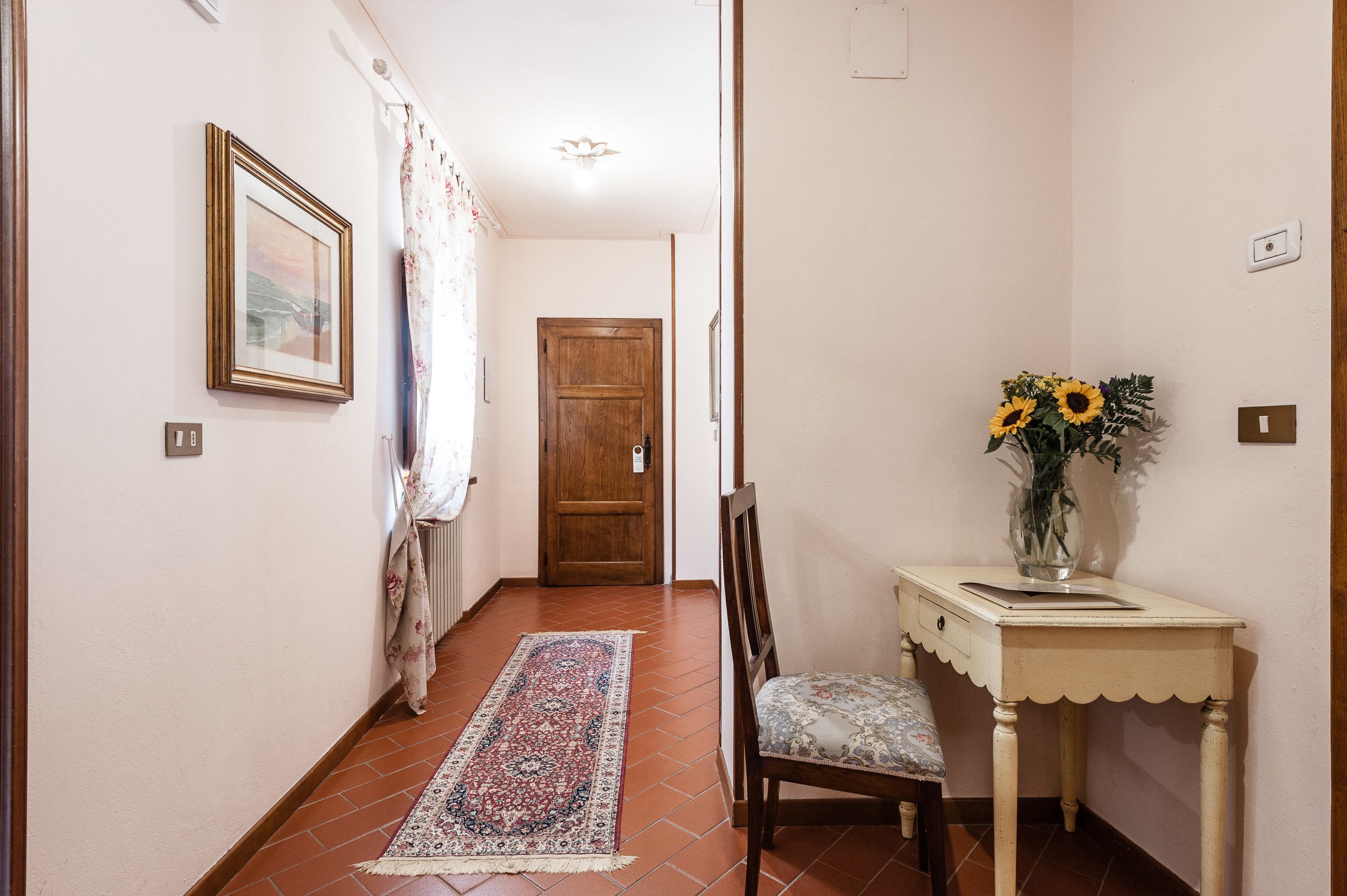 A hallway with a wooden door, a chair, a small table with a vase of sunflowers, and a patterned rug.