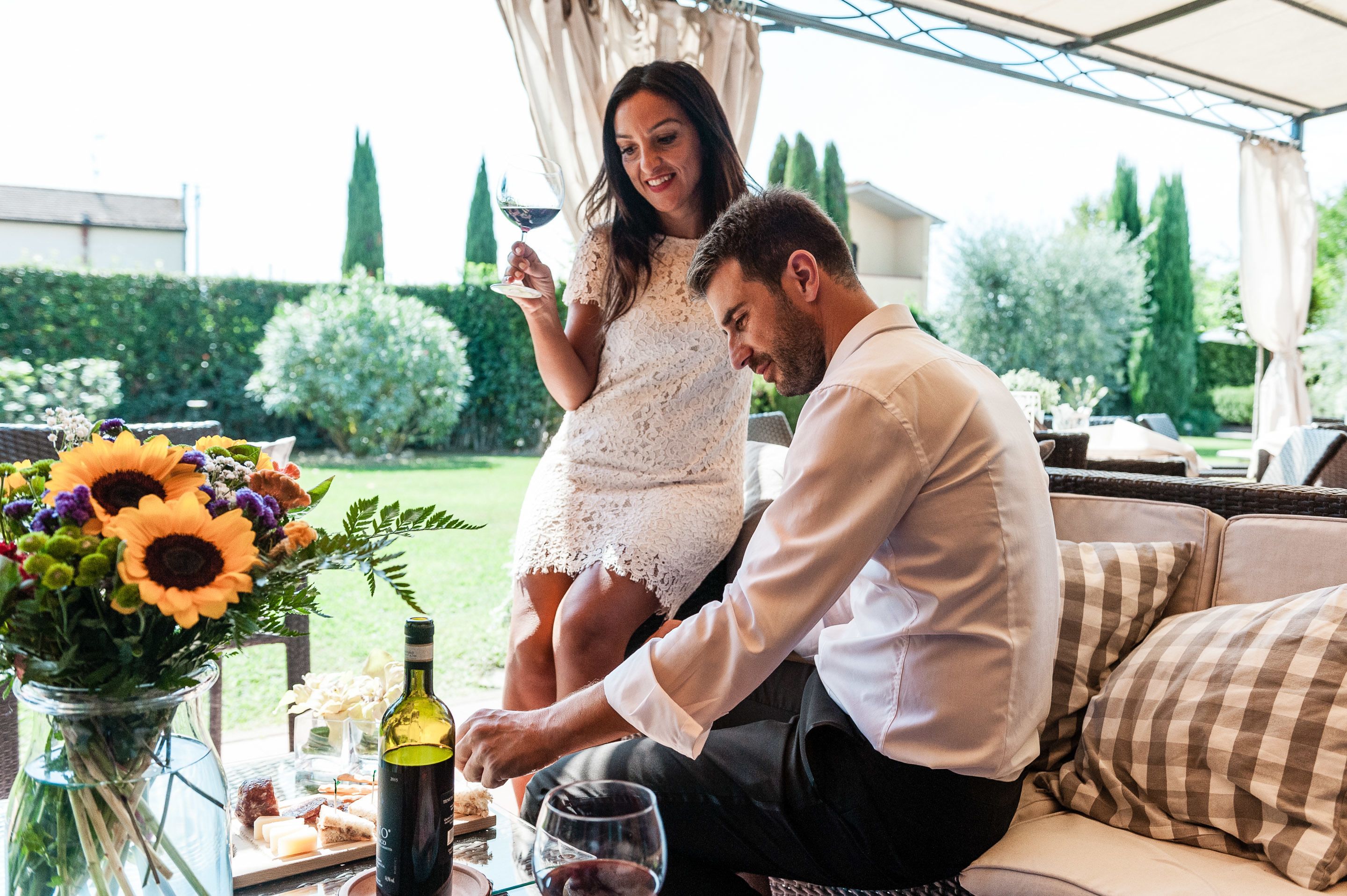 A couple seated at an outdoor table with a bottle of wine, glasses, and a floral arrangement.