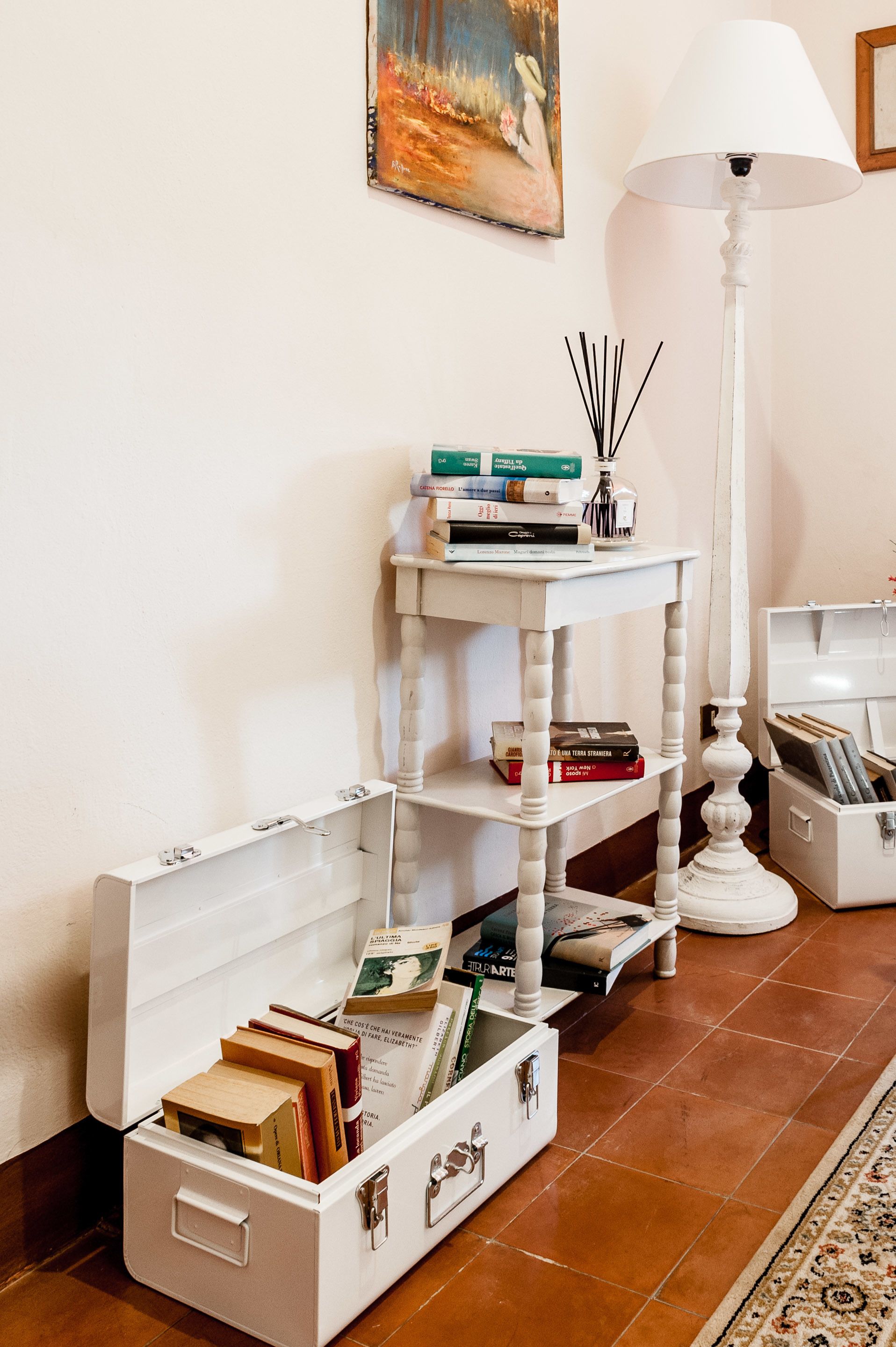 A white table with books and a lamp, a white cabinet with drawers, and a painting on the wall.