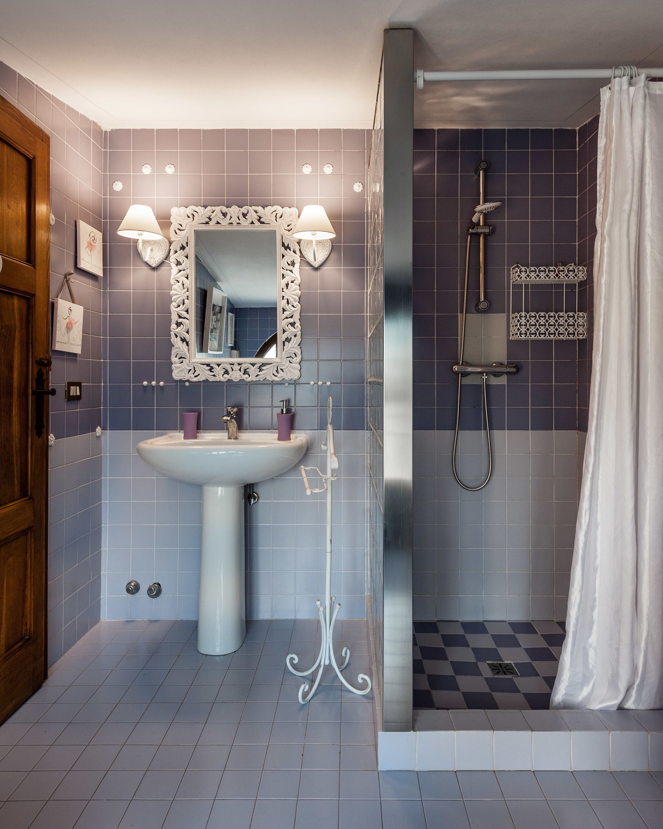 Une salle de bain avec un lavabo sur colonne, un grand miroir encadré de deux appliques murales, une douche avec un rideau et un carrelage à motifs à carreaux.