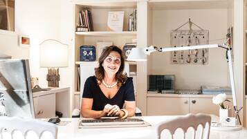 Una mujer sentada en un escritorio con una computadora, rodeada de libros y una lámpara.
