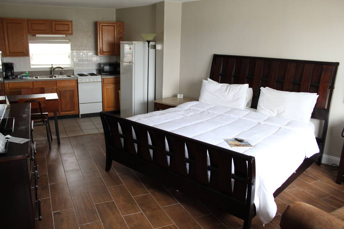 A bedroom with a large bed, a desk with a chair, a kitchen area with cabinets and appliances, and a bathroom with a toilet and sink.