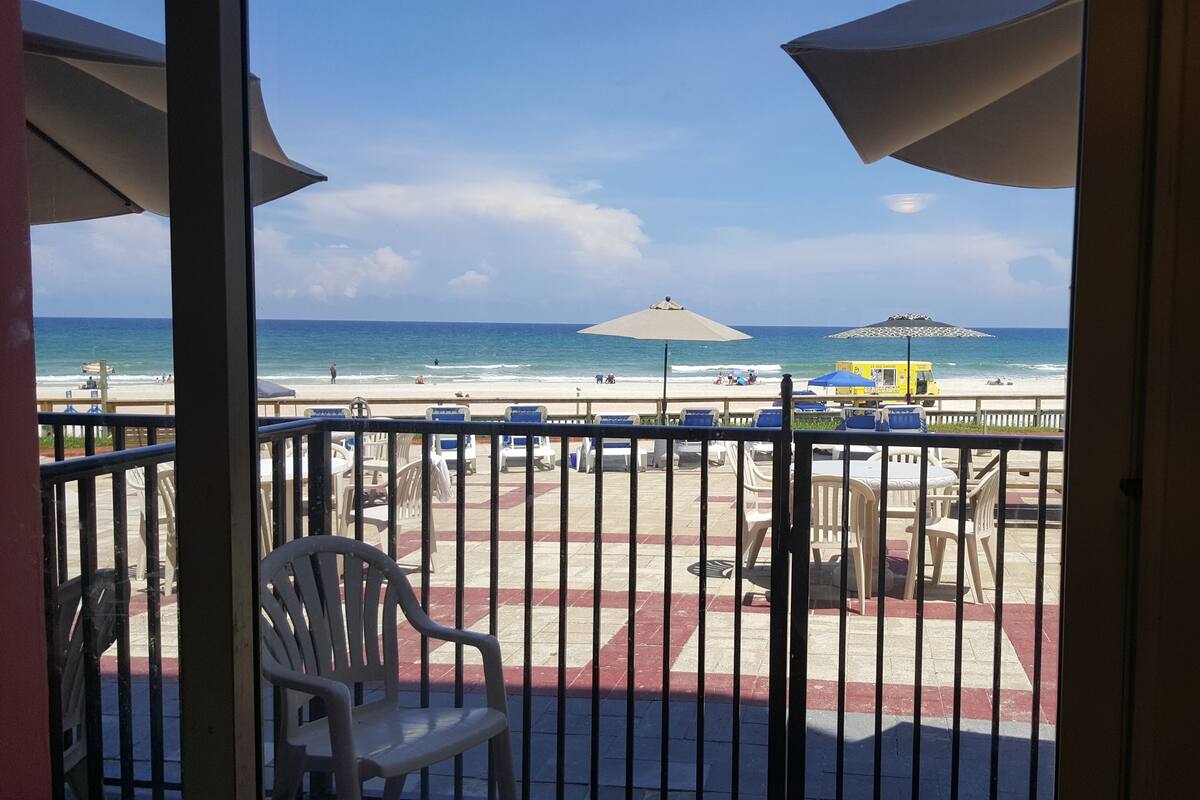 A balcony with a view of the beach, featuring outdoor seating and umbrellas.