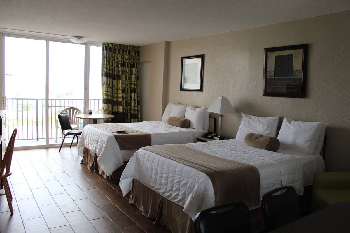 A hotel room with two beds, a balcony, a table, and chairs.