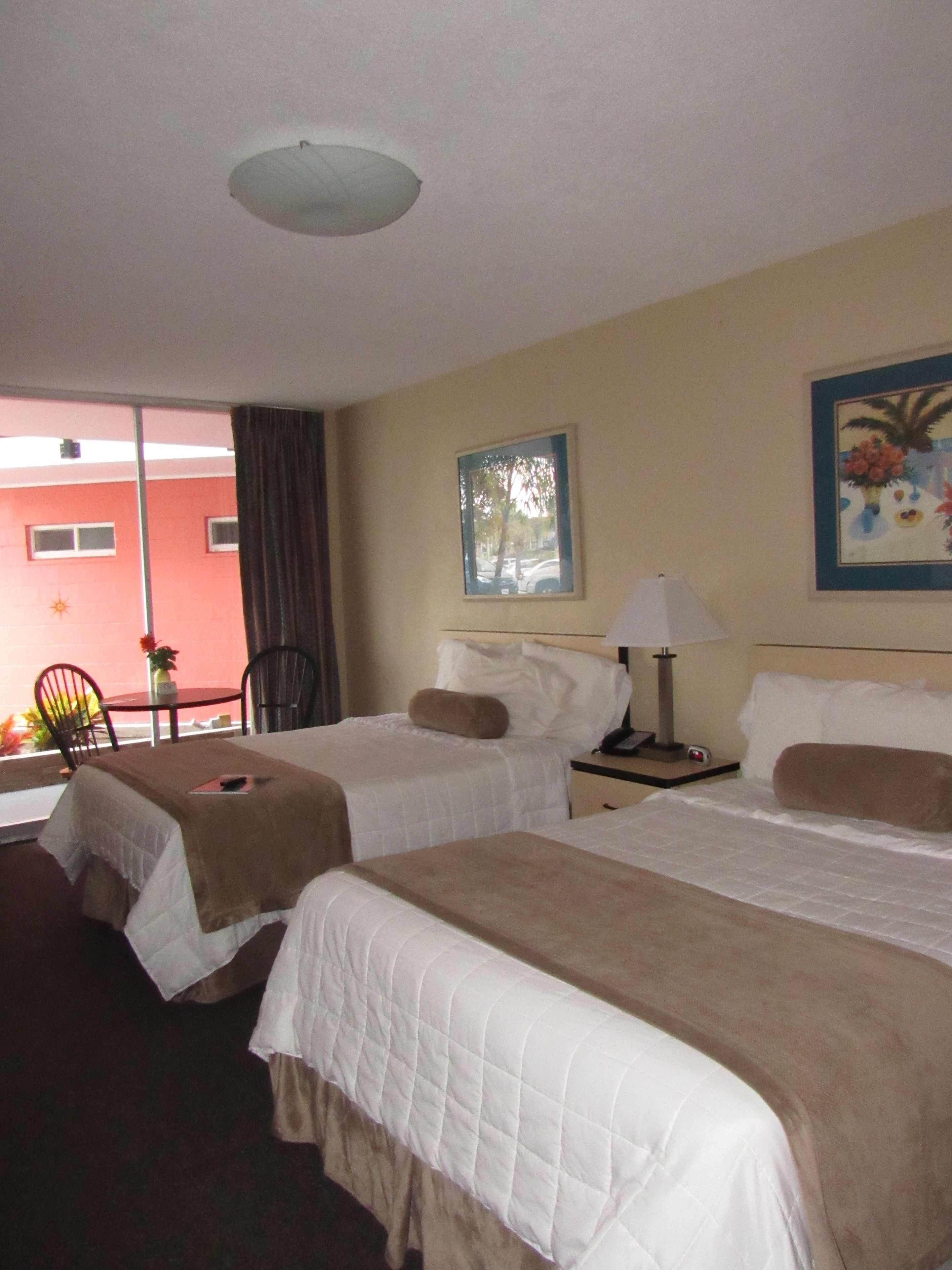 A hotel room with two beds, a small table, a chair, a window, and a painting on the wall.