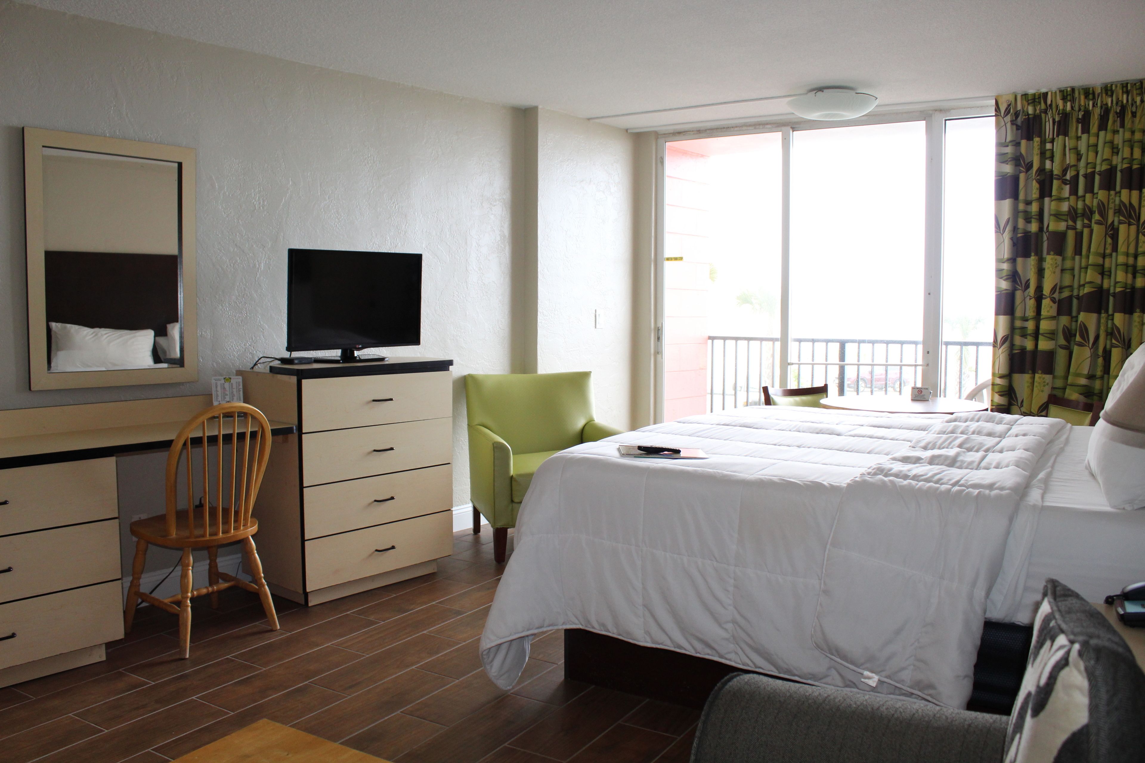 Habitación de hotel con cama, escritorio con televisor, una silla, cómoda y ventana con balcón.