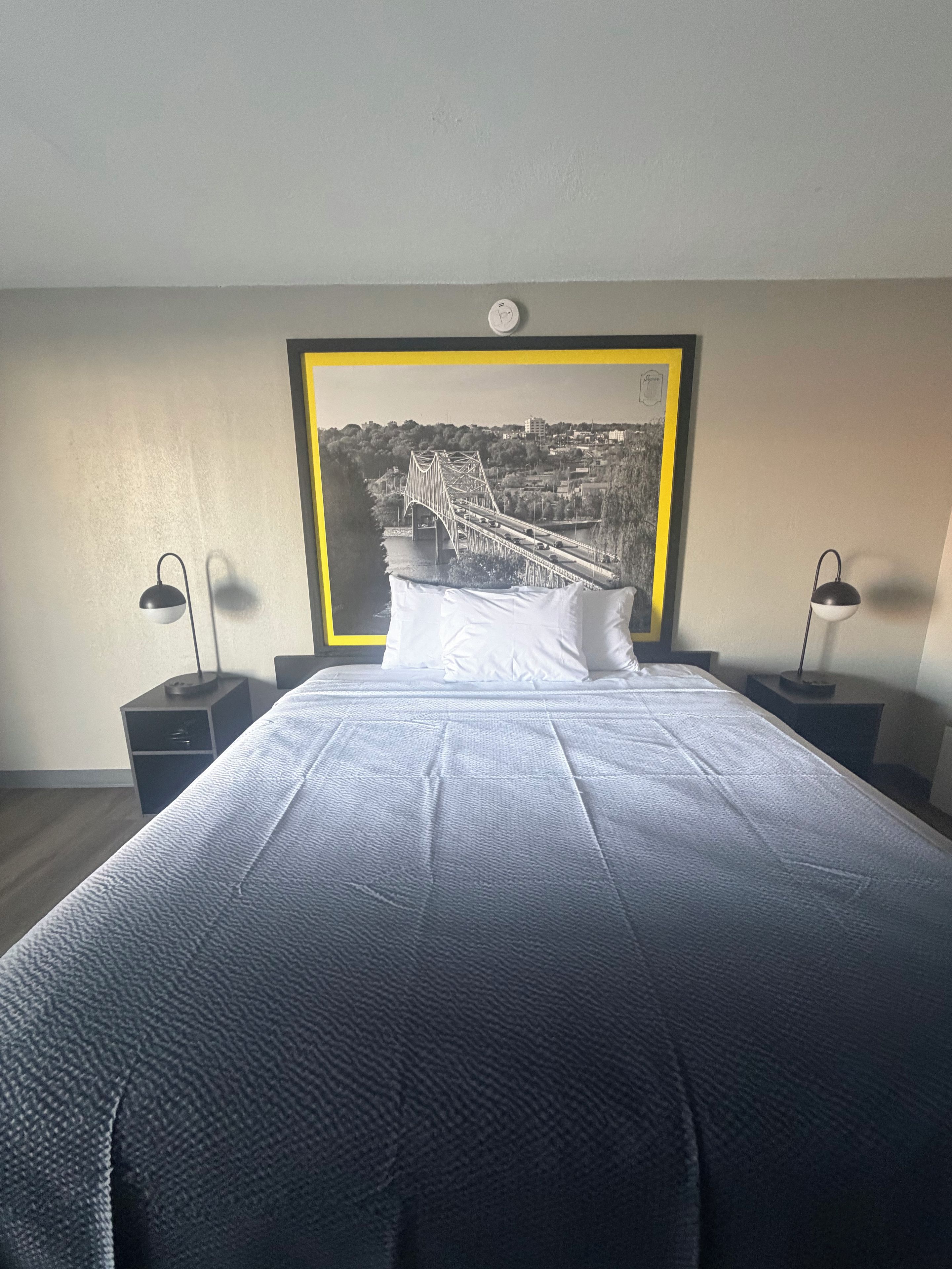 Une chambre d’hôtel avec un grand lit, deux tables de chevet, un tableau encadré représentant un pont et une horloge au mur.