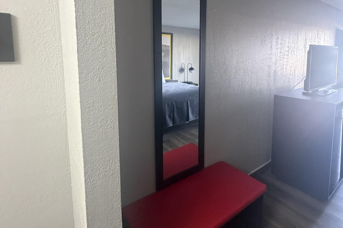 A hotel room with a bed, a desk, a mirror, and a red bench.
