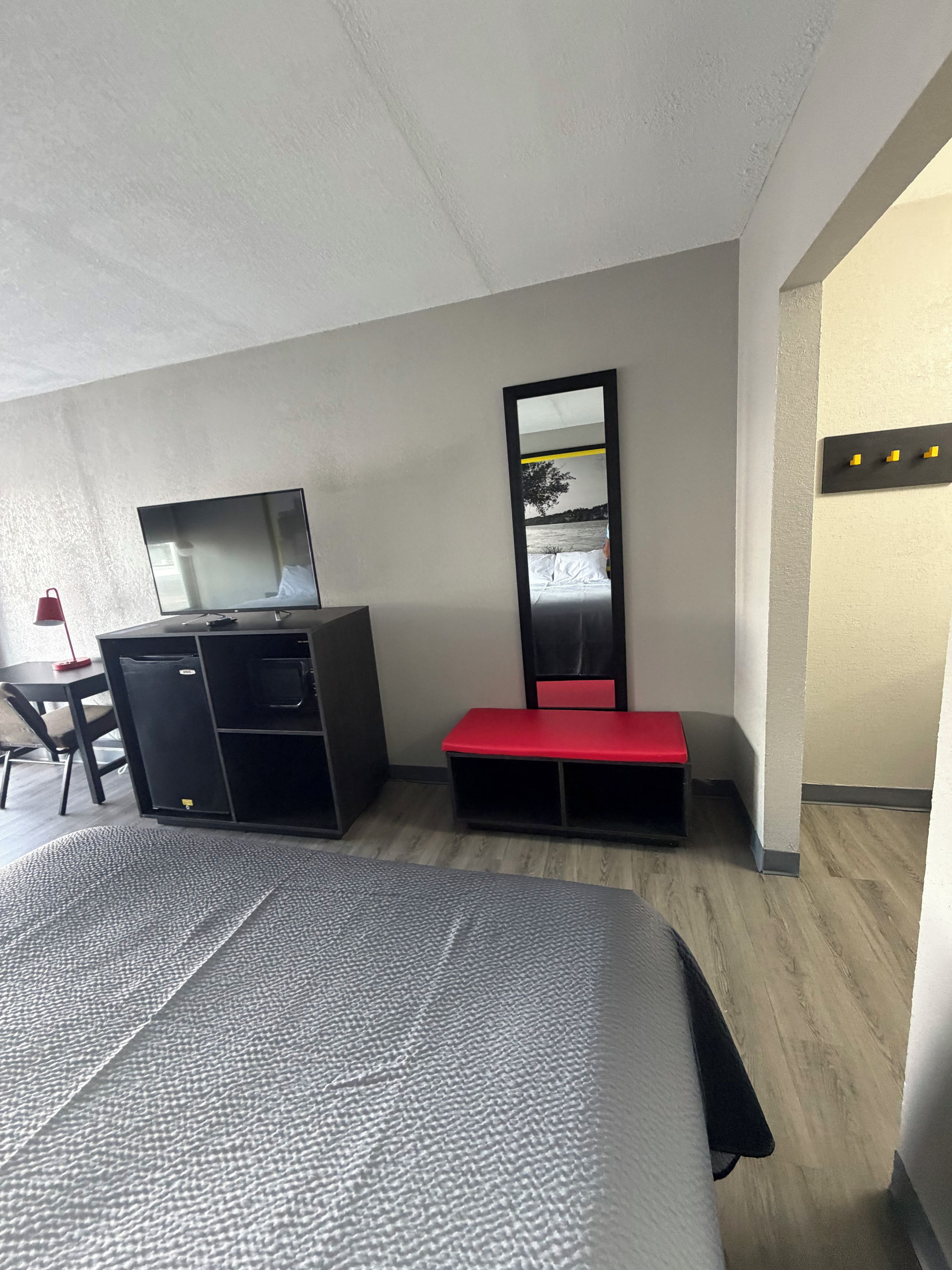 A hotel room with a bed, a desk with a computer, a mirror, and a red bench.