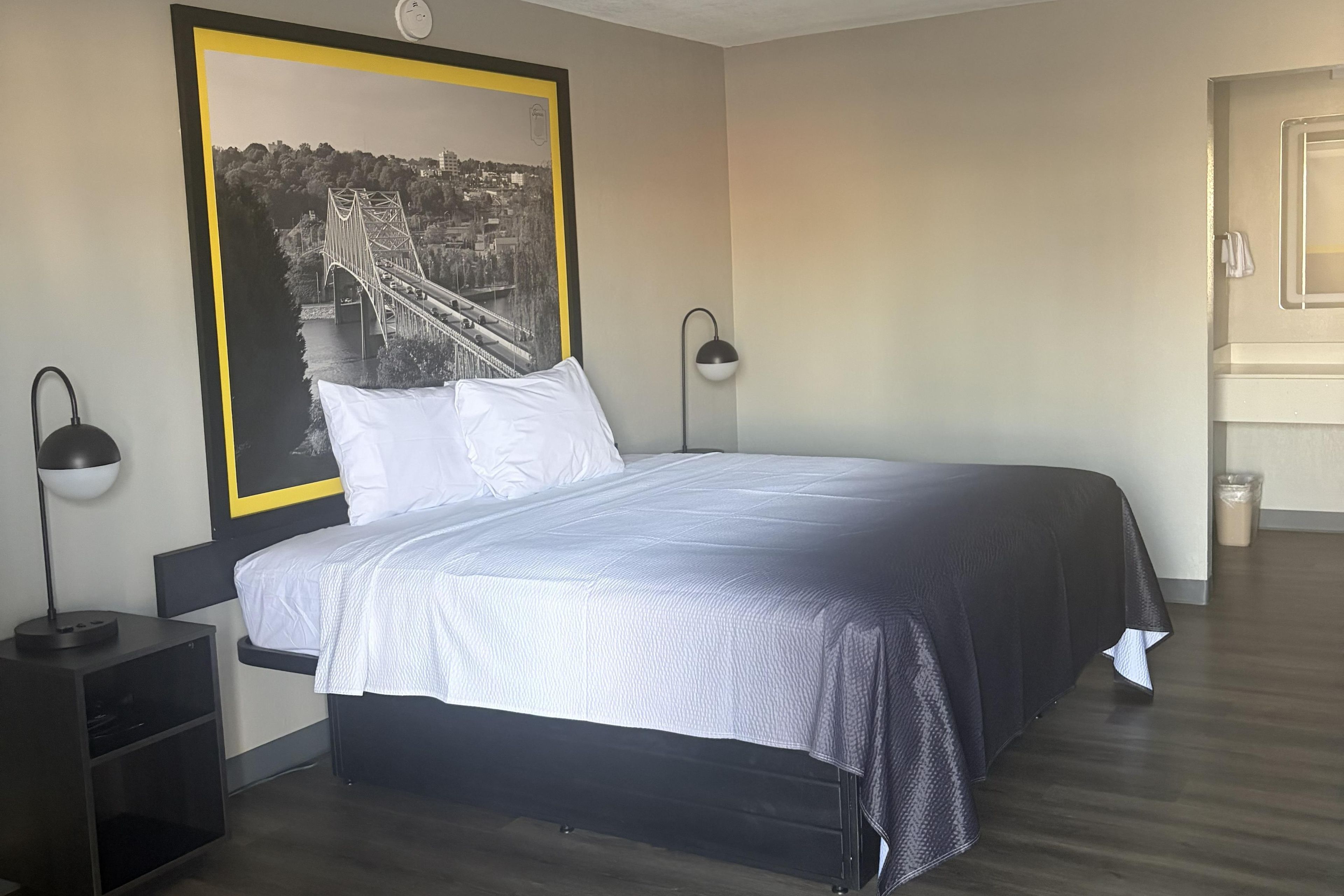 Un dormitorio con una cama, dos lámparas de noche, una mesita de noche, un cuadro grande enmarcado en la pared y una puerta a la derecha.