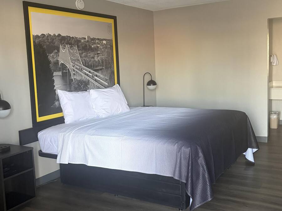 Un dormitorio con una cama, dos lámparas de noche, una mesita de noche, un cuadro grande enmarcado en la pared y una puerta a la derecha.