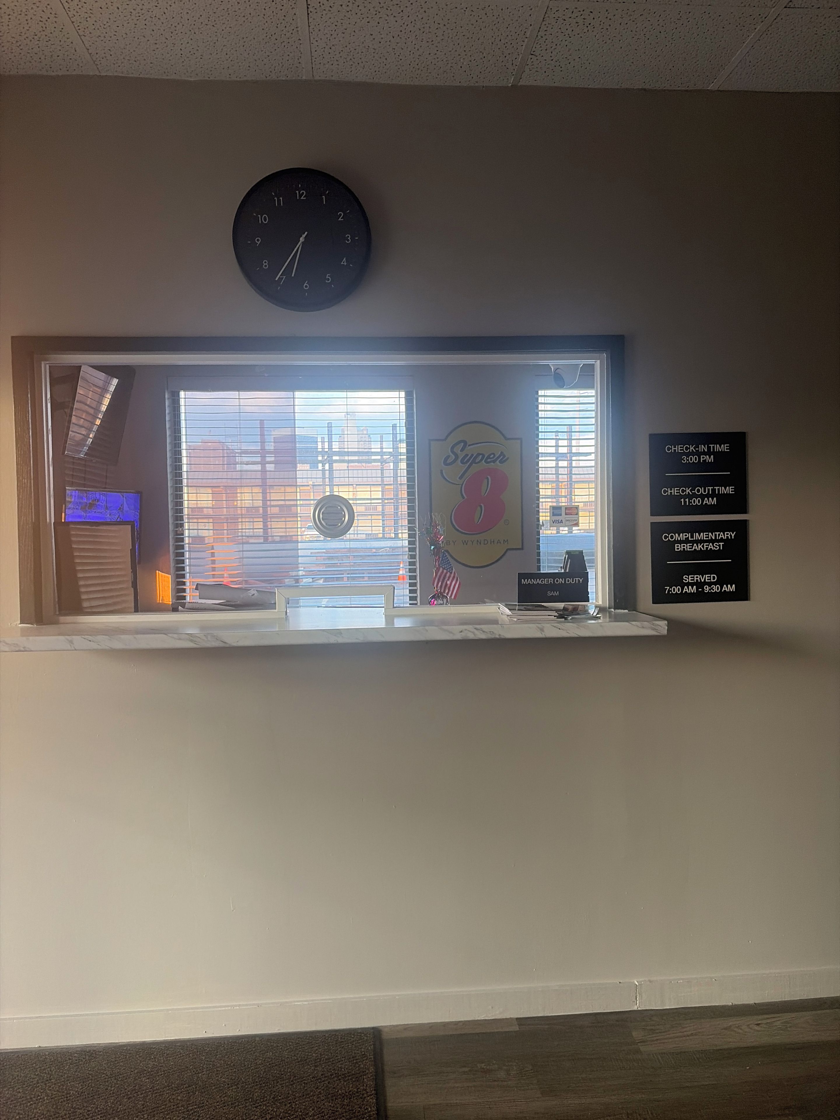 Zona de recepción de un hotel con un reloj, una ventana con persianas y una pared que muestra los horarios de registro de entrada/salida y detalles del desayuno de cortesía.