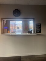 Zona de recepción de un hotel con un reloj, una ventana con persianas y una pared que muestra los horarios de registro de entrada/salida y detalles del desayuno de cortesía.