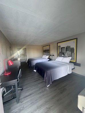 A hotel room with two beds, a desk with a chair, a red lamp, and a cityscape painting on the wall.