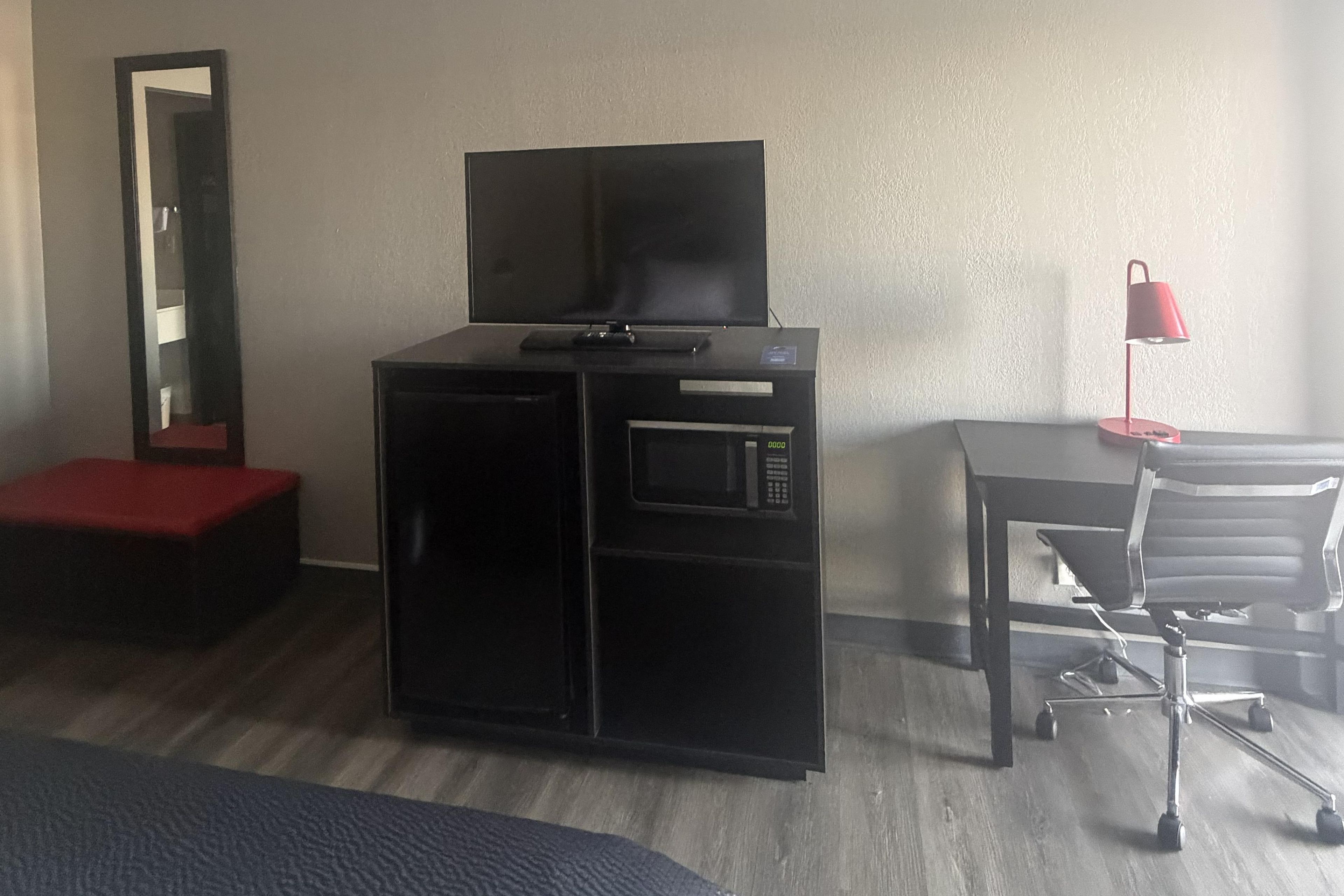 A hotel room with a bed, a desk with a chair, a television on a cabinet, and a red lamp.