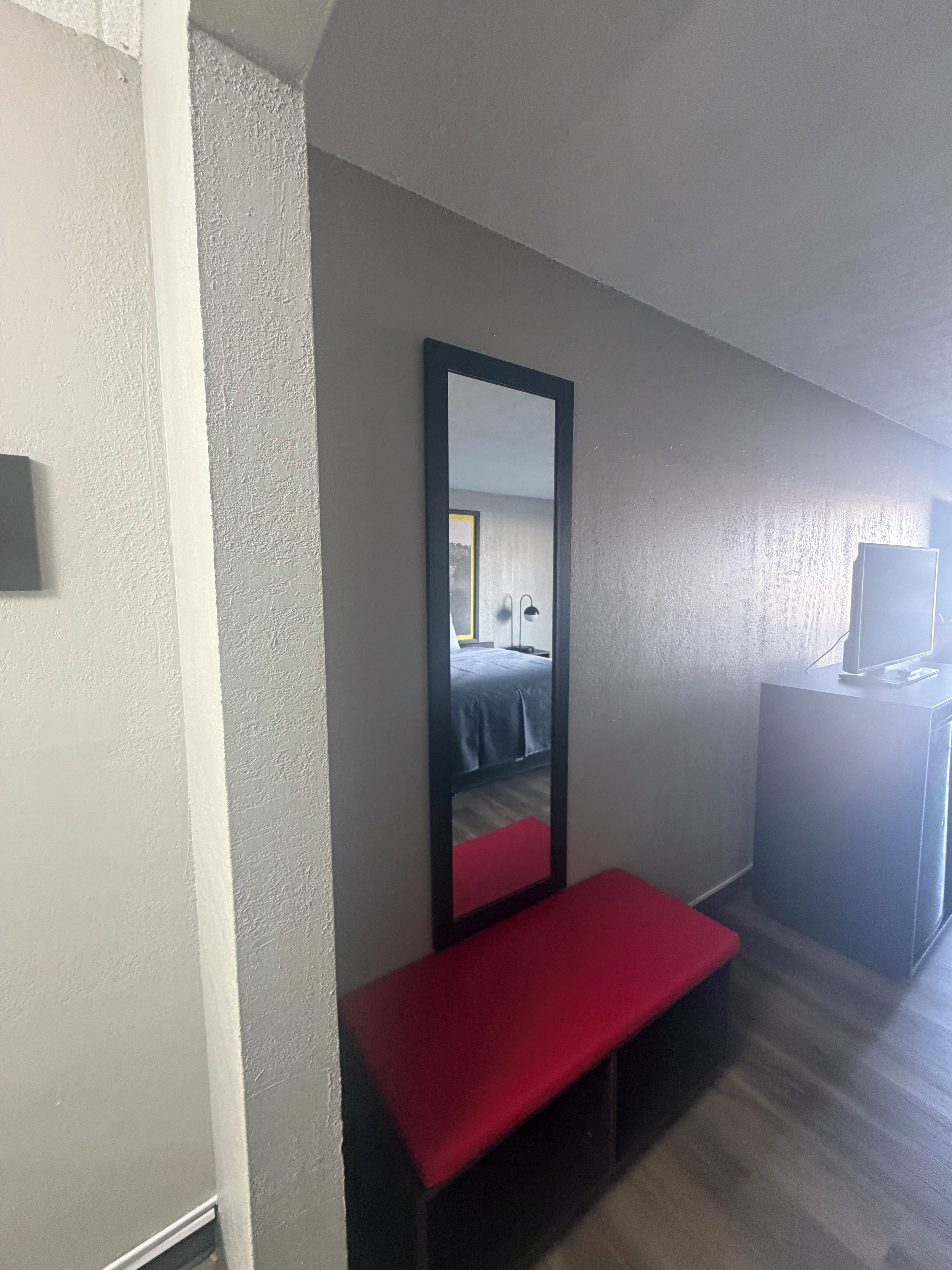 Una habitación de hotel con una cama, un banco rojo, un espejo grande y un televisor sobre un mueble.