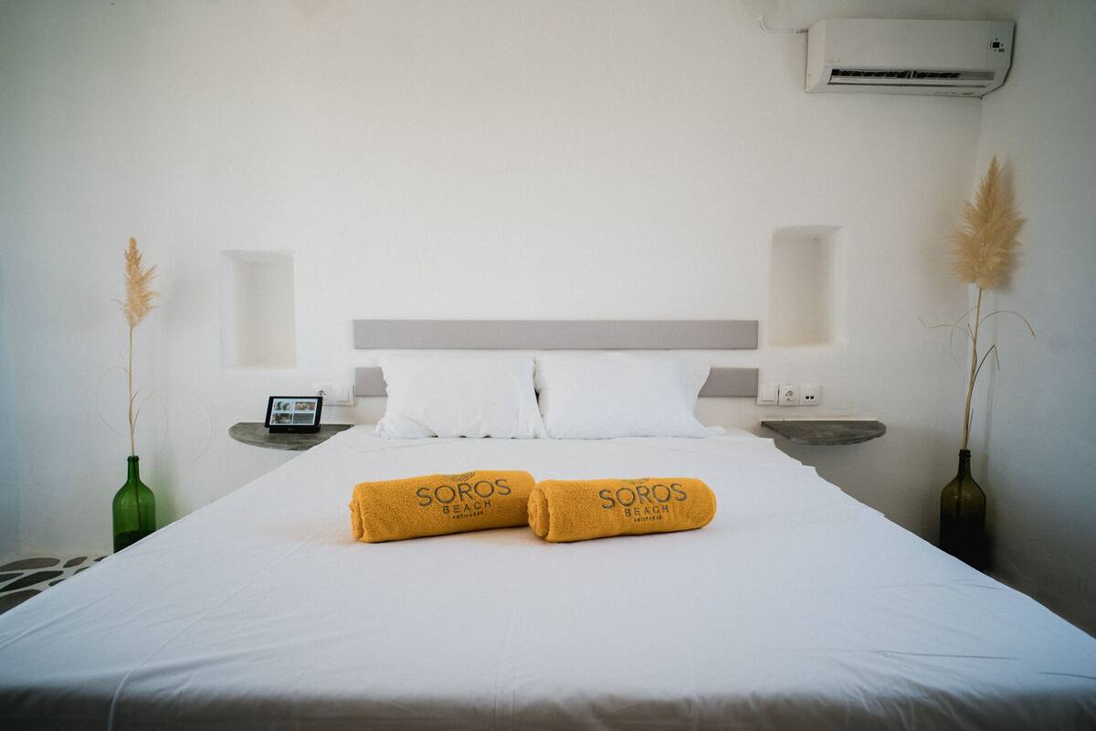 Una cama bien hecha con dos toallas con la etiqueta "Soros Beach" y una mesita de noche con un teléfono y un pequeño marco de fotos.