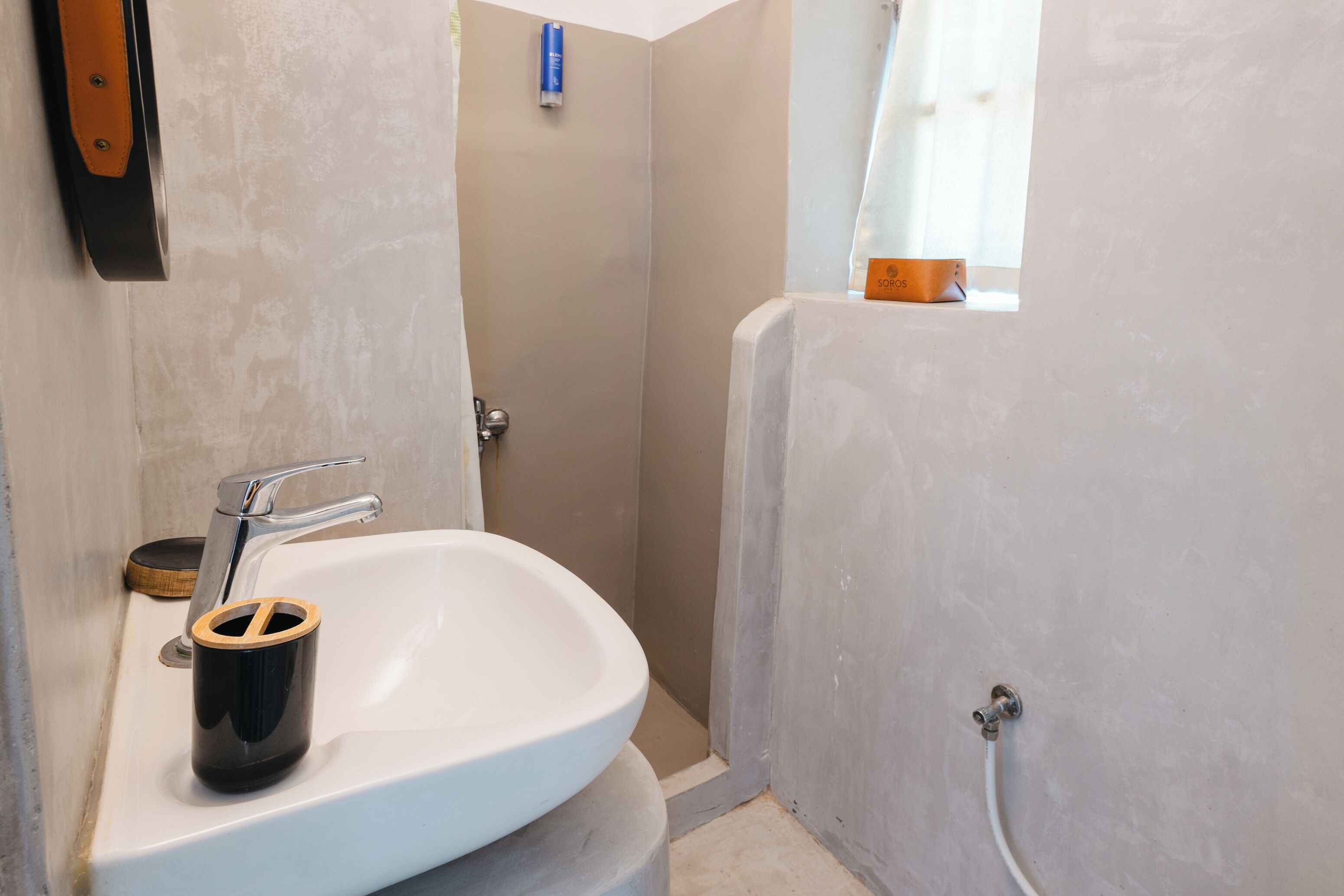 Un baño con un lavamanos blanco, un cubo de basura negro, una zona de ducha y una ventana con cortina.