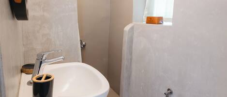 Un baño con un lavamanos blanco, un cubo de basura negro, una zona de ducha y una ventana con cortina.