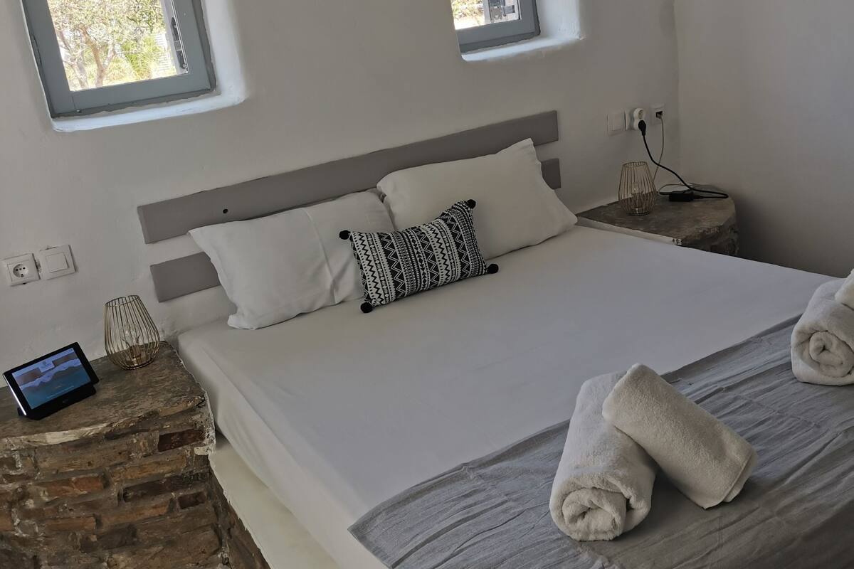 Un dormitorio con una cama, dos ventanas, una mesita de noche con lámpara y una tableta.