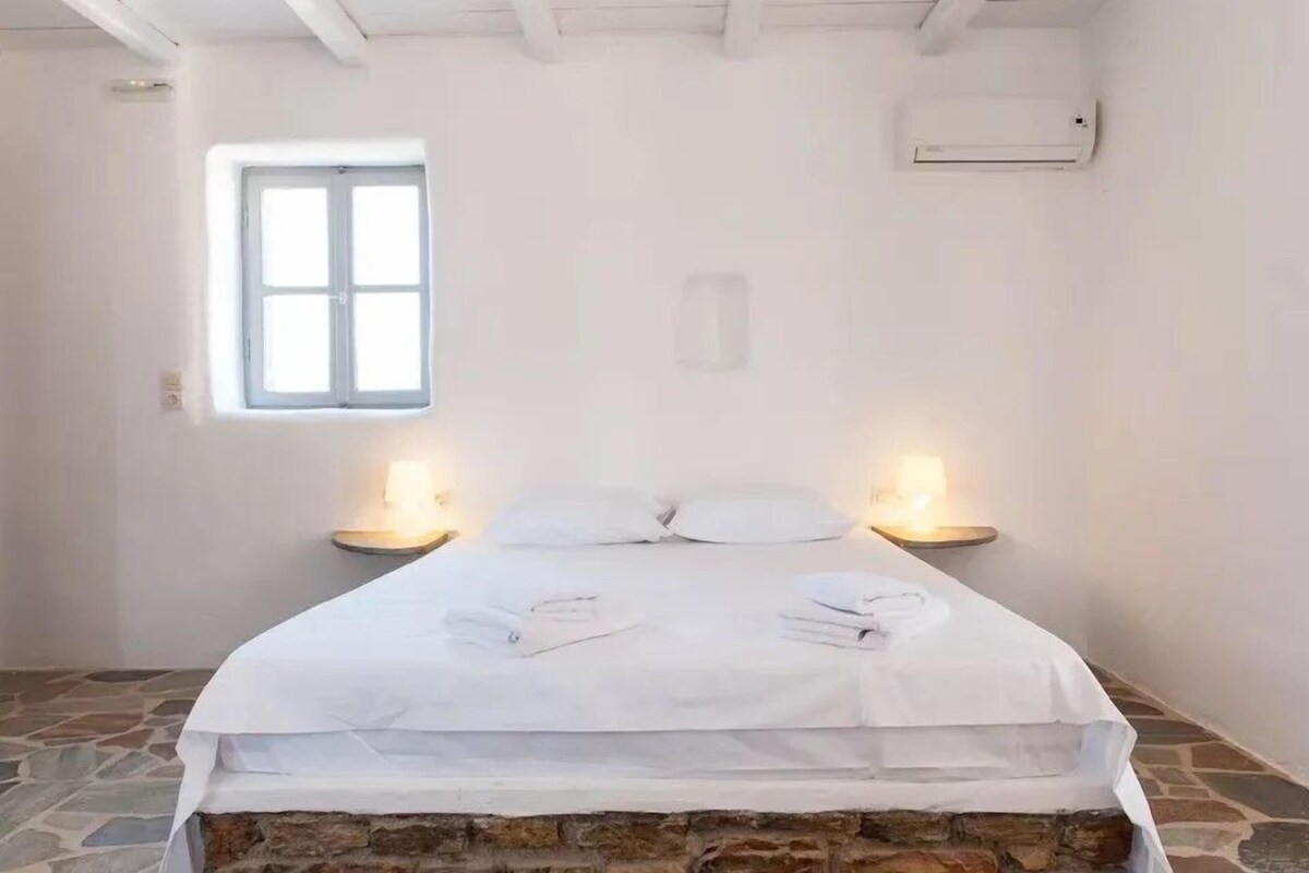 A bedroom with a stone floor, a bed with white linens, two bedside lamps, and a window.