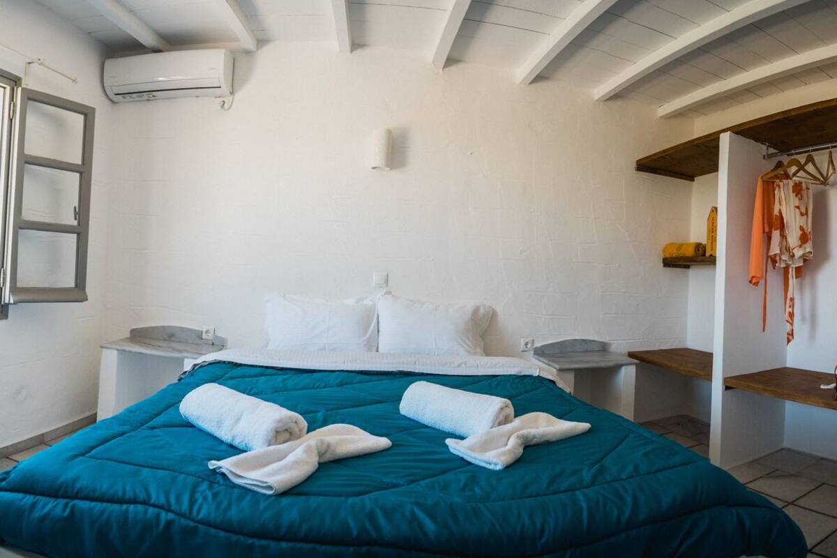 Un dormitorio con colcha color esmeralda, almohadas blancas, un banco de madera y una ventana con contraventanas abiertas.