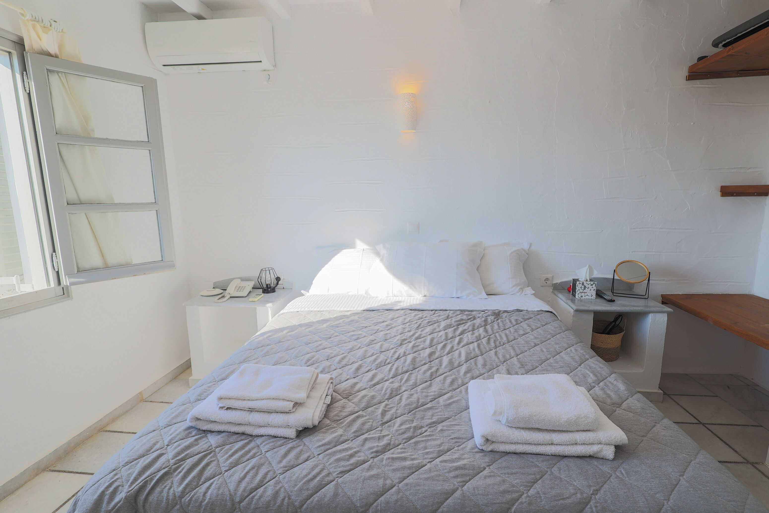 A hotel room with a bed, bedside table, a lamp, a window, and a wall-mounted air conditioner.