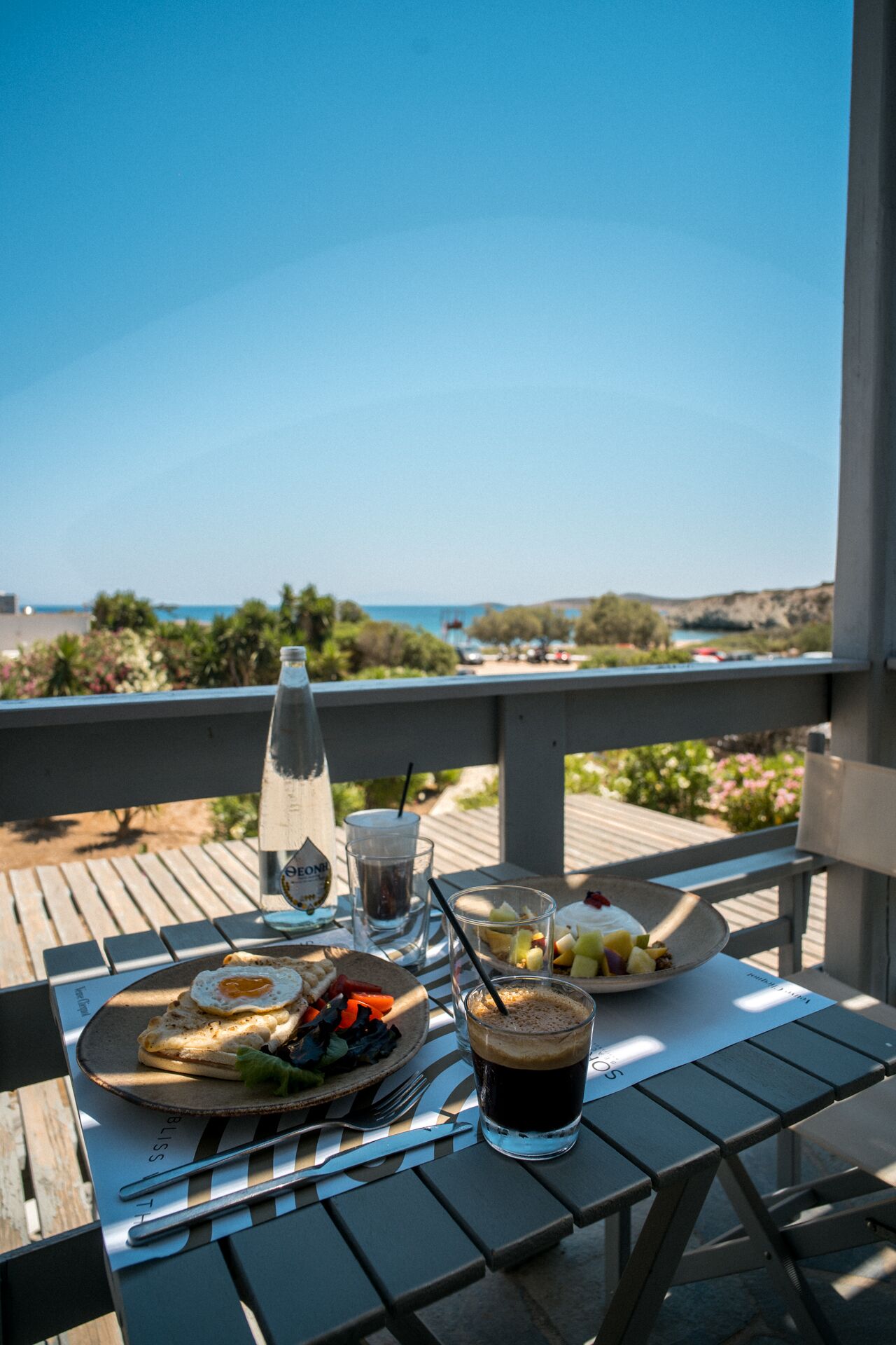 A table with breakfast items, a bottle, and a glass of beverage on a wooden deck with a view of the ocean.