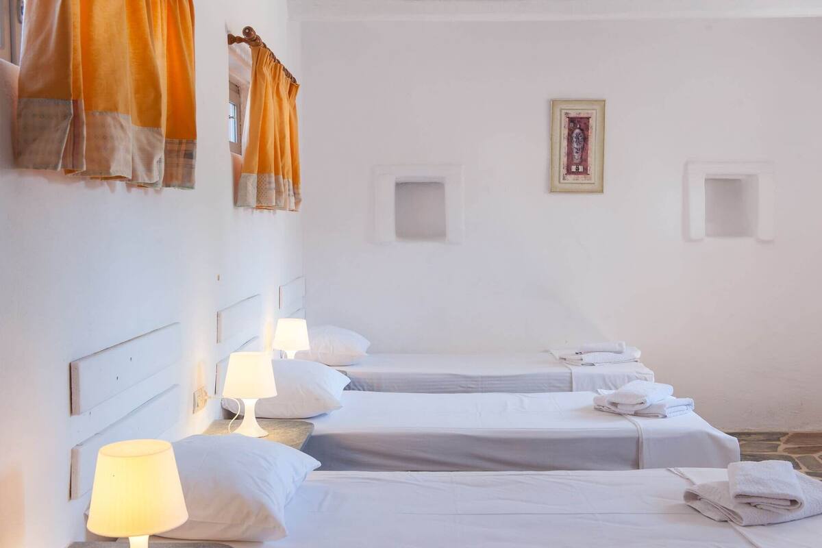 Habitación de hotel con tres camas, cada una con ropa blanca y almohadas, y cabeceros. Hay dos lámparas de pared en cada cama.