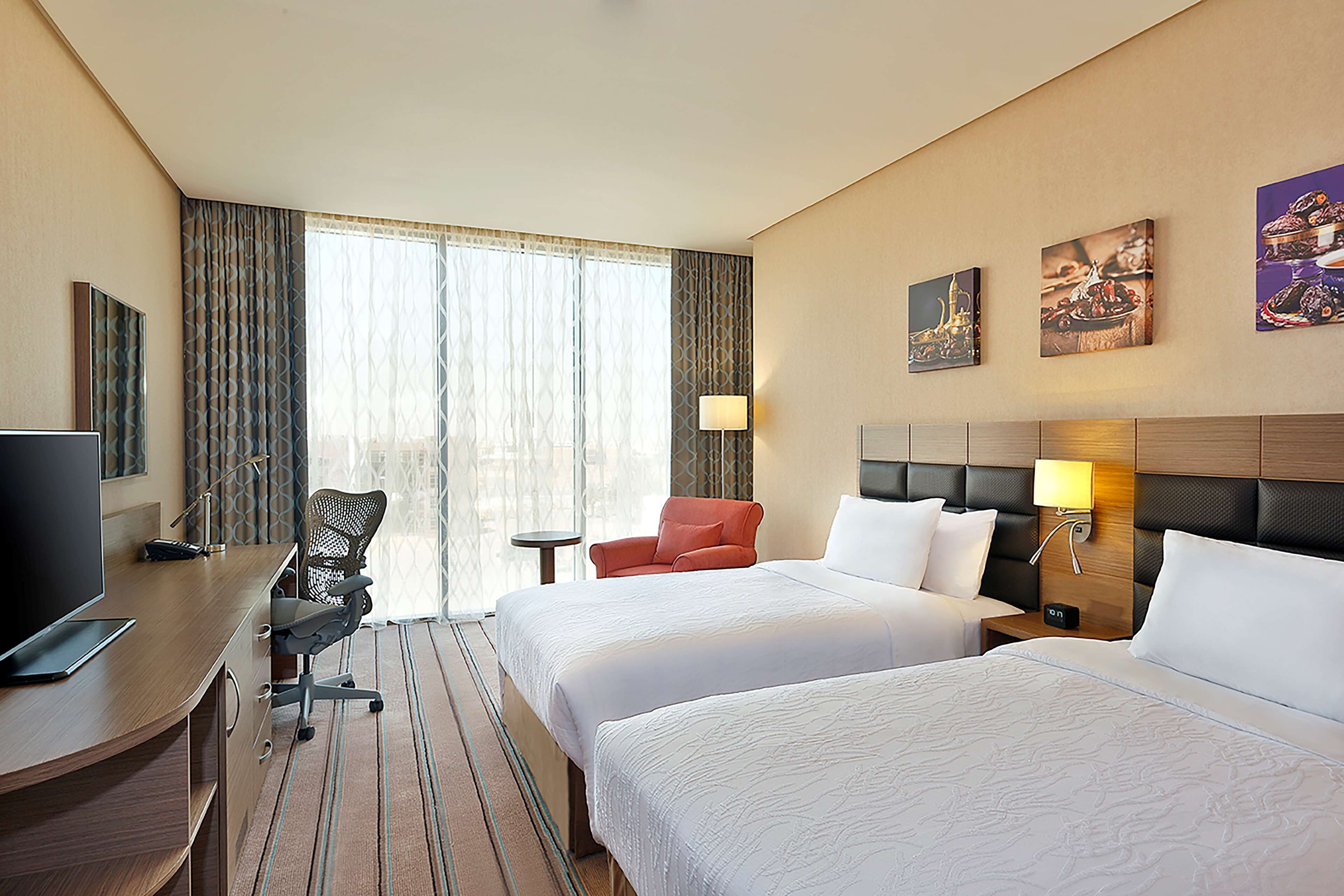 A hotel room with two beds, a desk with a laptop, a chair, a TV, and a window with curtains.