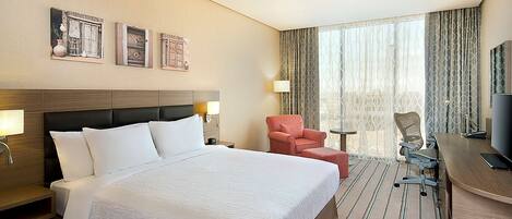 Quarto de hotel com uma cama grande, uma escrivaninha com cadeira, televisão e janela com cortinas.