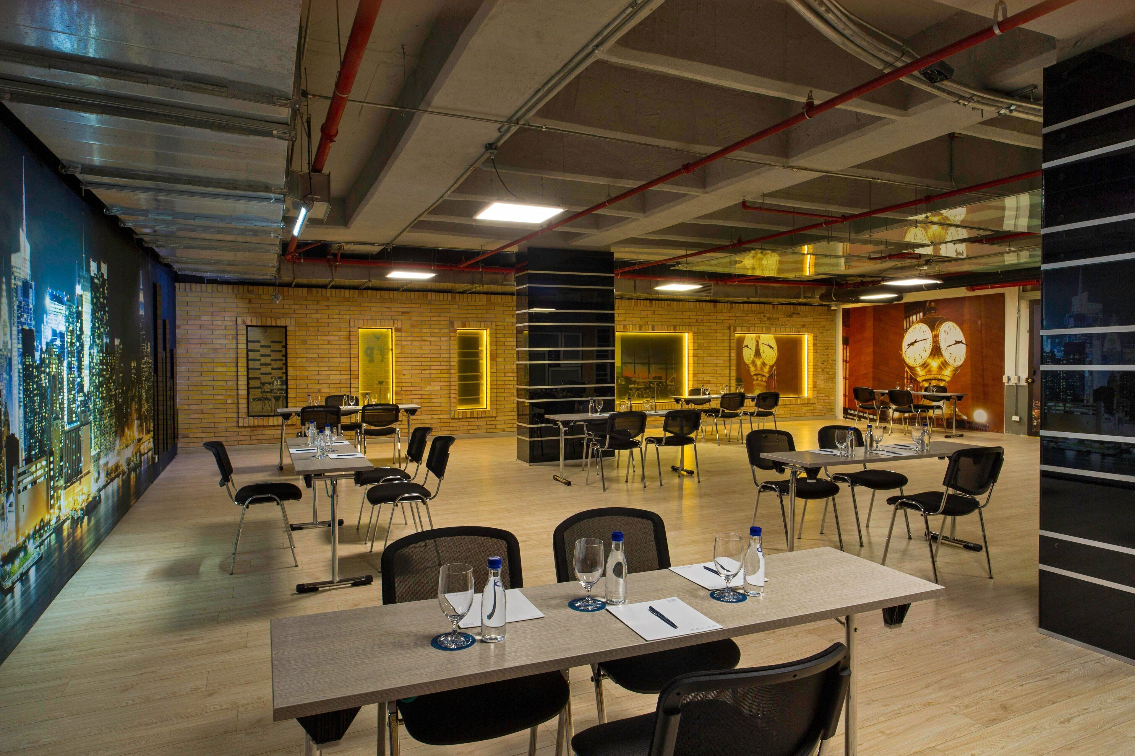 A spacious conference room with multiple tables and chairs, equipped with water bottles and glasses, and a cityscape mural on the wall.
