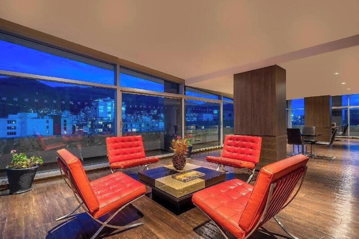 A modern interior with a seating area featuring red chairs, a central table, and large windows offering a city view.