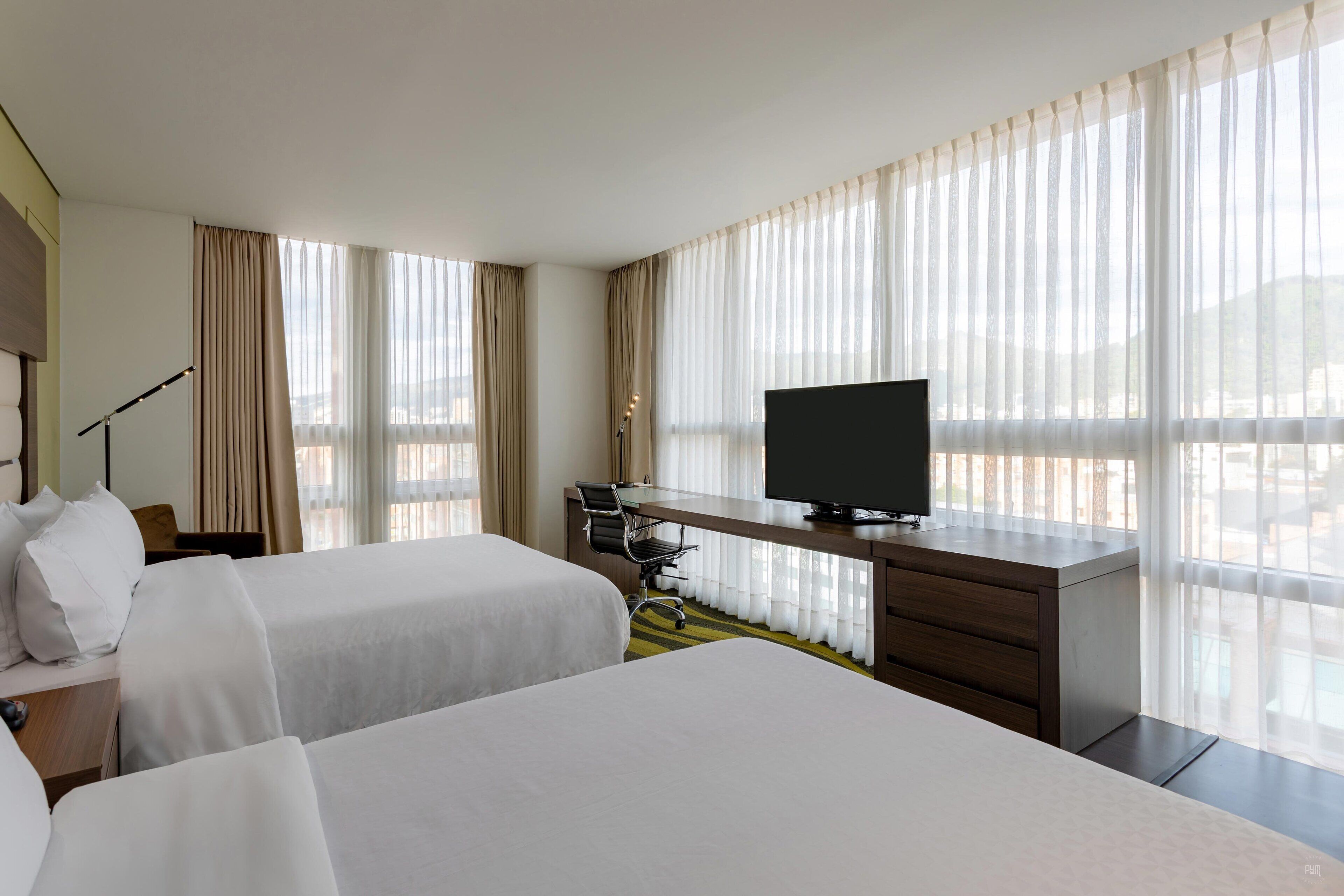 A hotel room with two beds, a desk with a television, and large windows with sheer curtains.